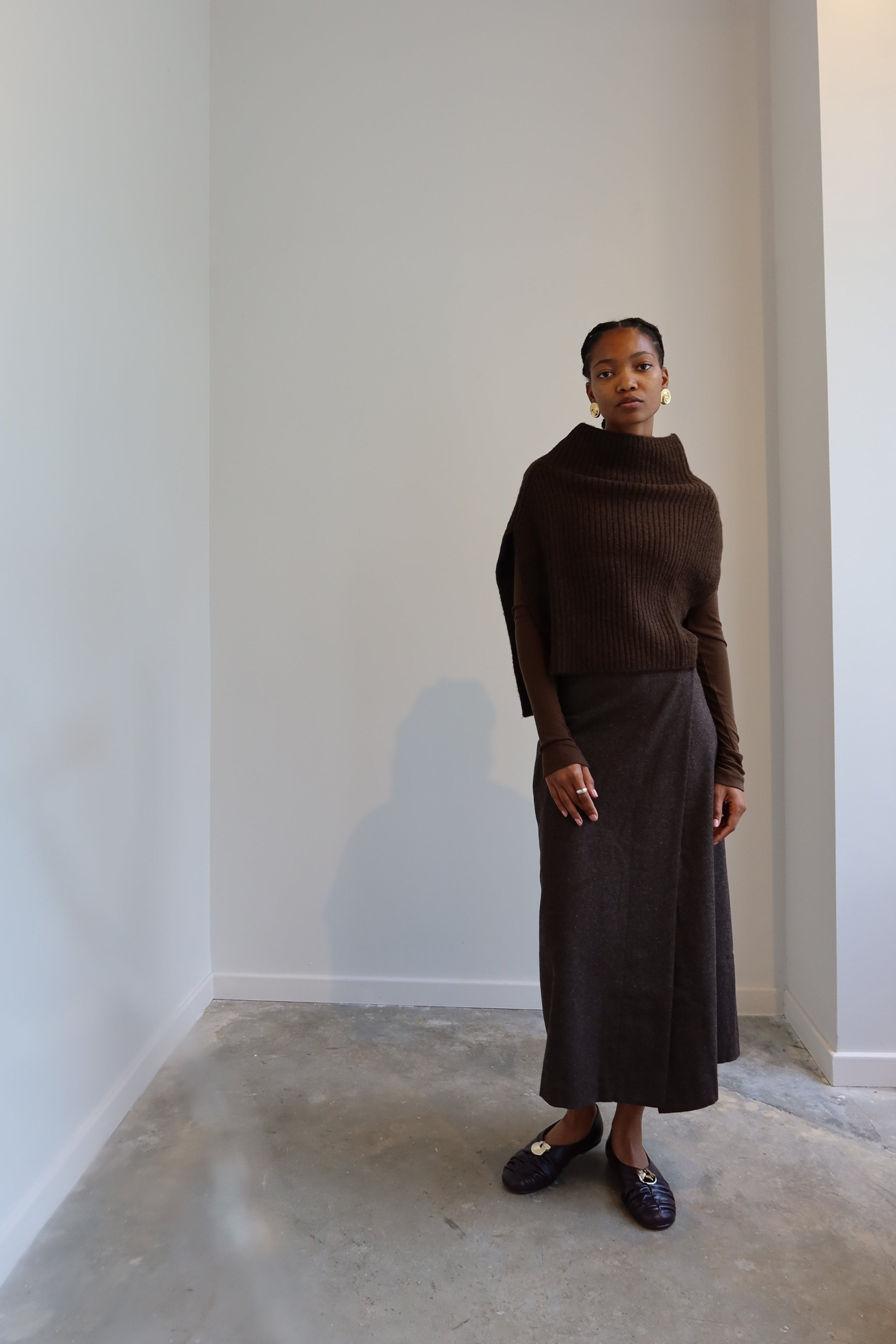 A person wearing an olive brown ribbed knitted Mijeong Park neck warmer with a mock neck neckline and tie waist detail, paired with a brown skirt at Alas Eius
