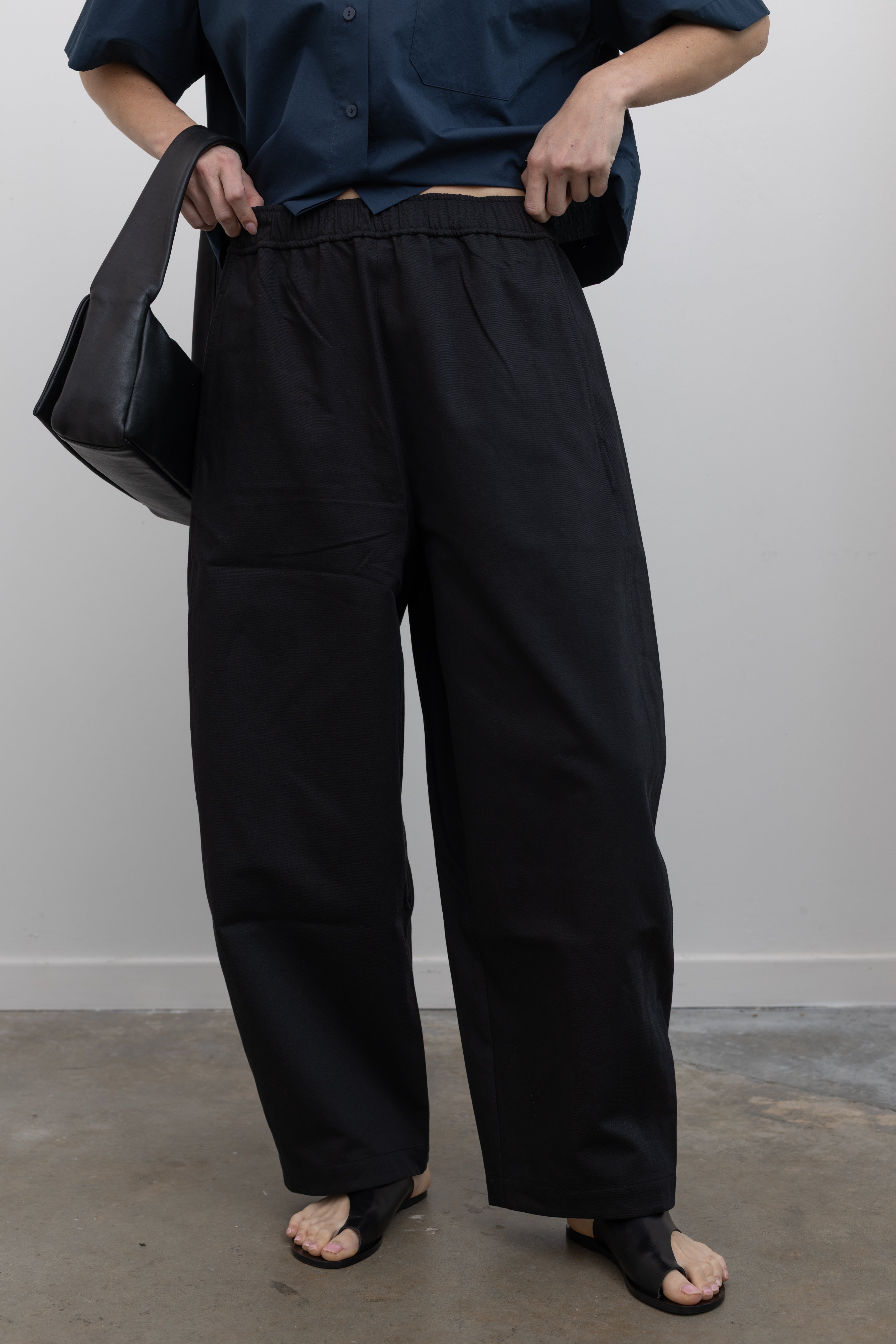 Close-up front view of contemporary womenswear spring fashion in Mijeong Park black baggy balloon pants with curved outseams, a relaxed fit, elastic waistband, and back patch pockets at an Alas Eius store. 