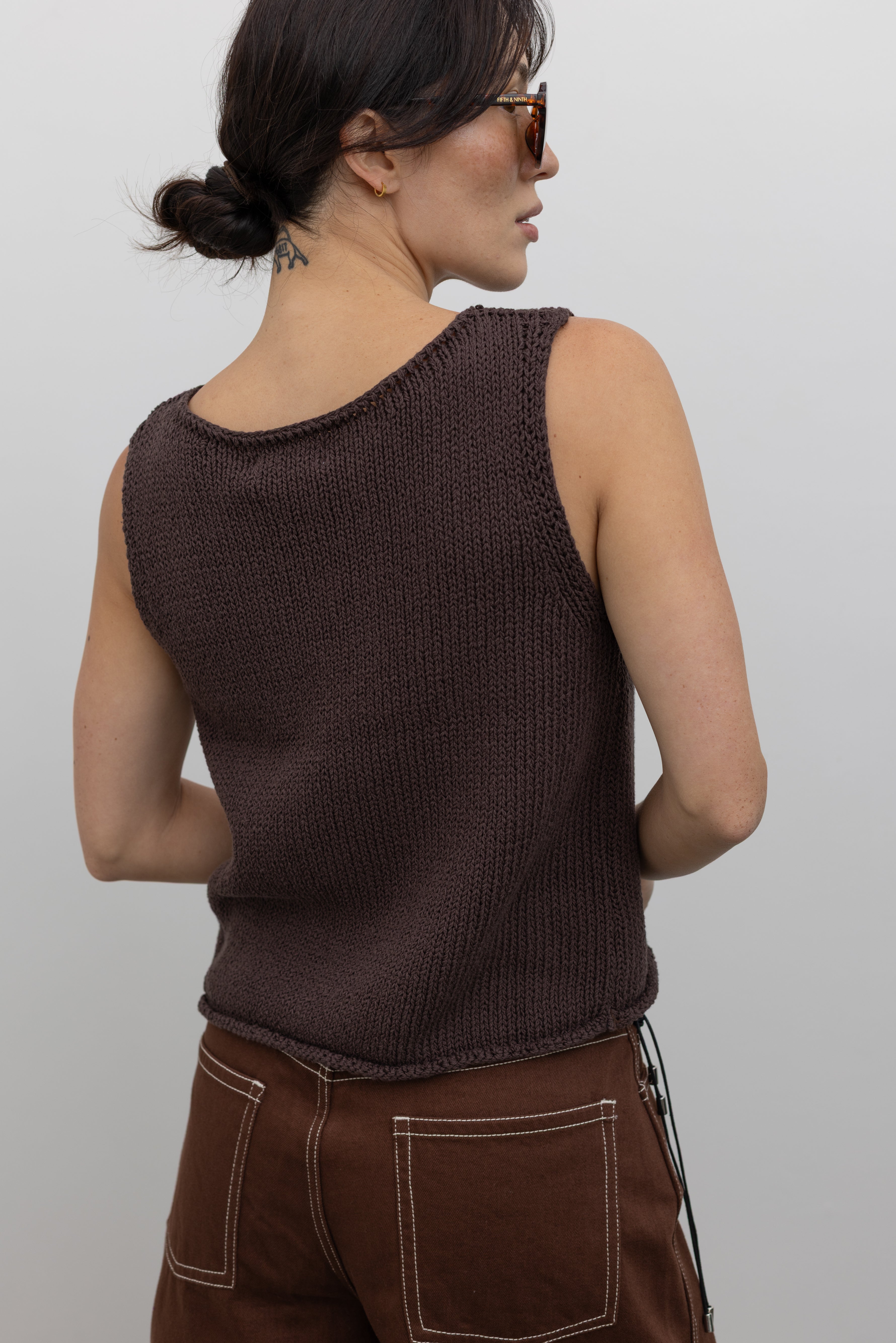 Close-up back view of a woman posing wearing modern minimalist spring and summer fashion in a Mijeong Park dark brown boat neck knit tank featuring a relaxed fit and rolled edges at the neck and hem paired with Kowtow Framework Organic Cotton Brown Denim Long Shorts that sit just above the knee and feature white contrast stitching at an Alas Eius store.  