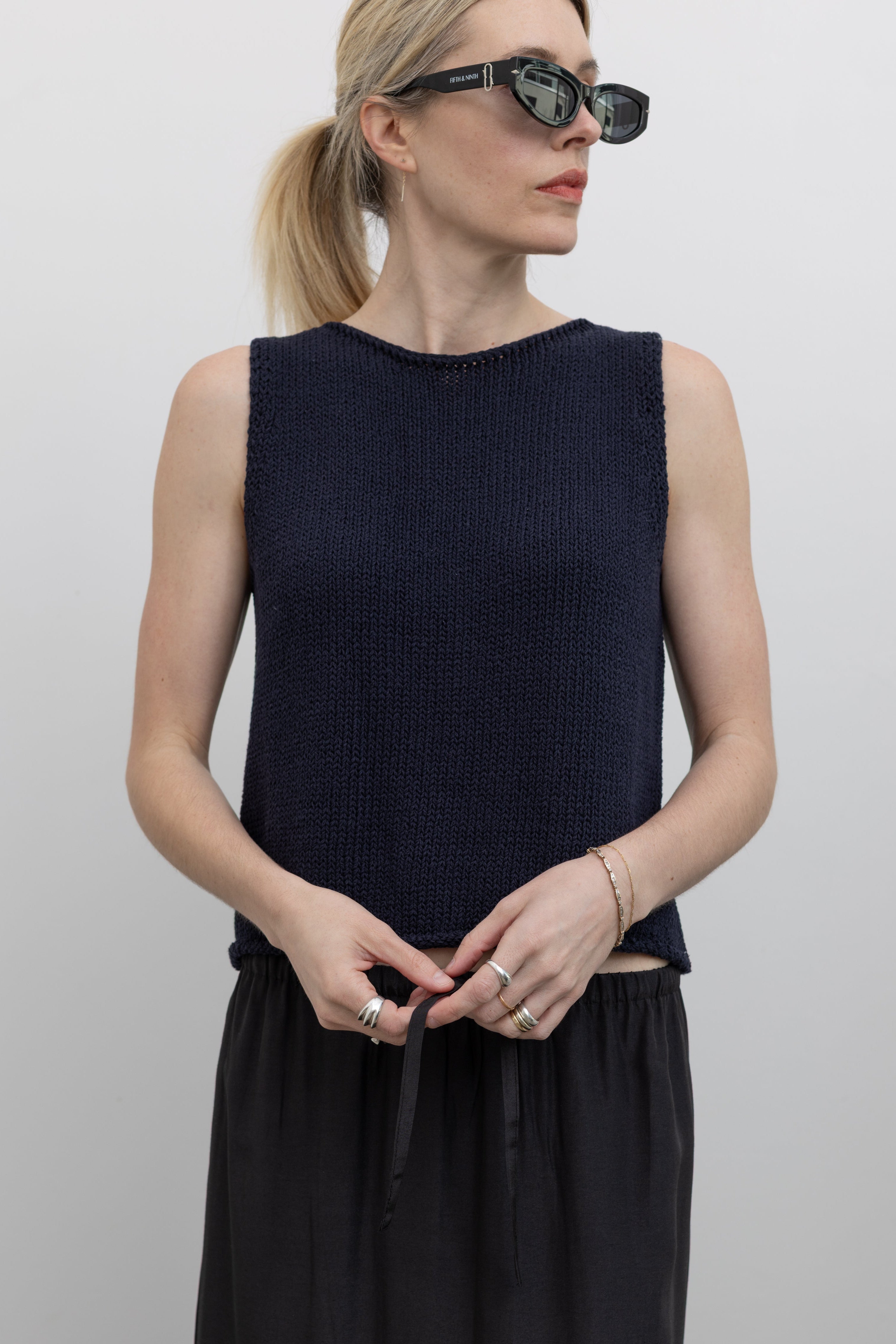 Close-up front view of a woman standing wearing spring fashion in a Mijeong Park navy blue boat neck knit sweater tank featuring rolled edges around the neckline and hem at an Alas Eius store. 