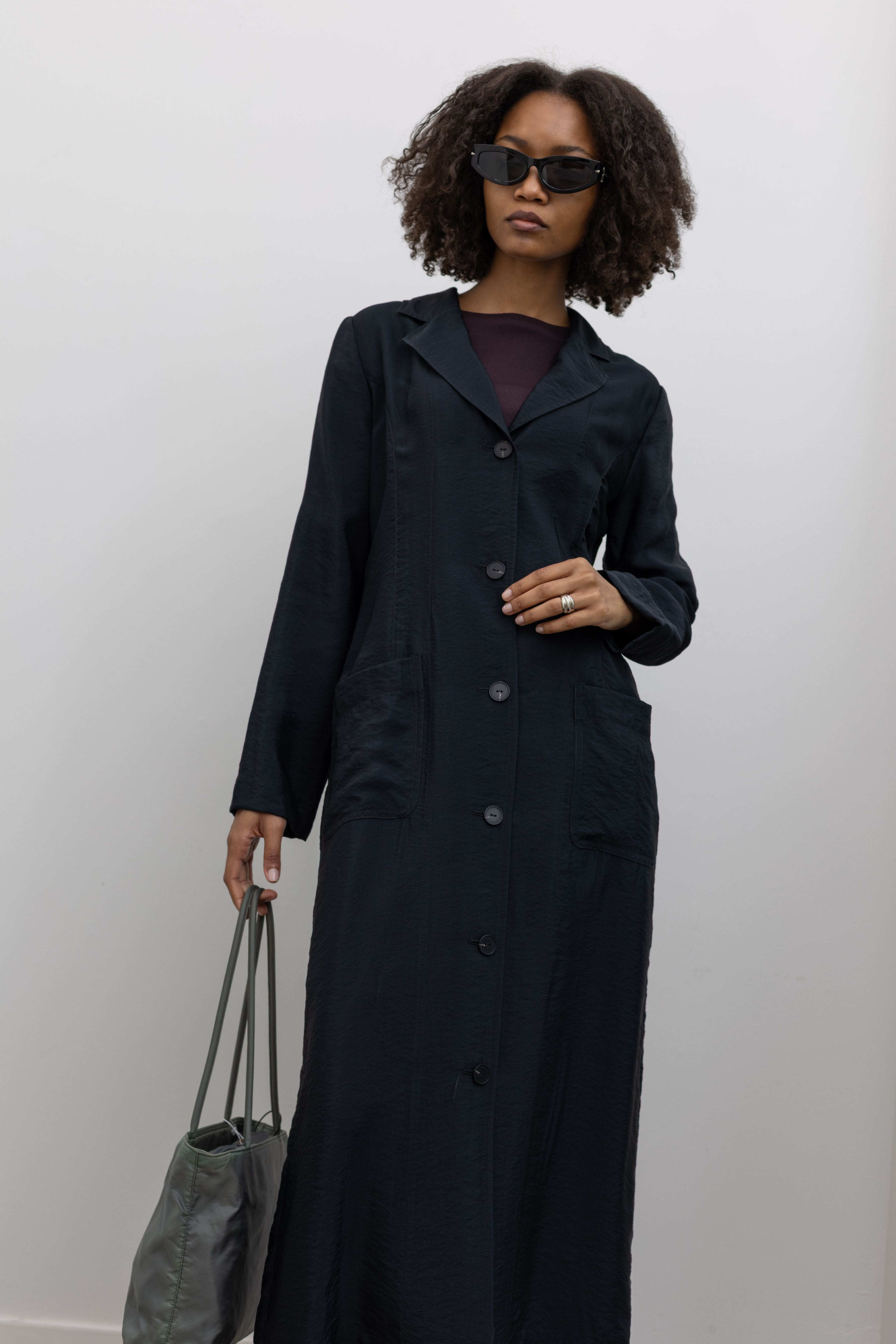 Full length front view of a woman in motion wearing sophisticated spring and summer womenswear fashion in a elegant Nomia Black Long Unlined Relaxed Fit Duster Jacket with 6 front buttons and 2 patch pockets on front hip at an Alas Eius store. Made in New York from a beautiful Japanese Acetate blend with a washed vintage finish.