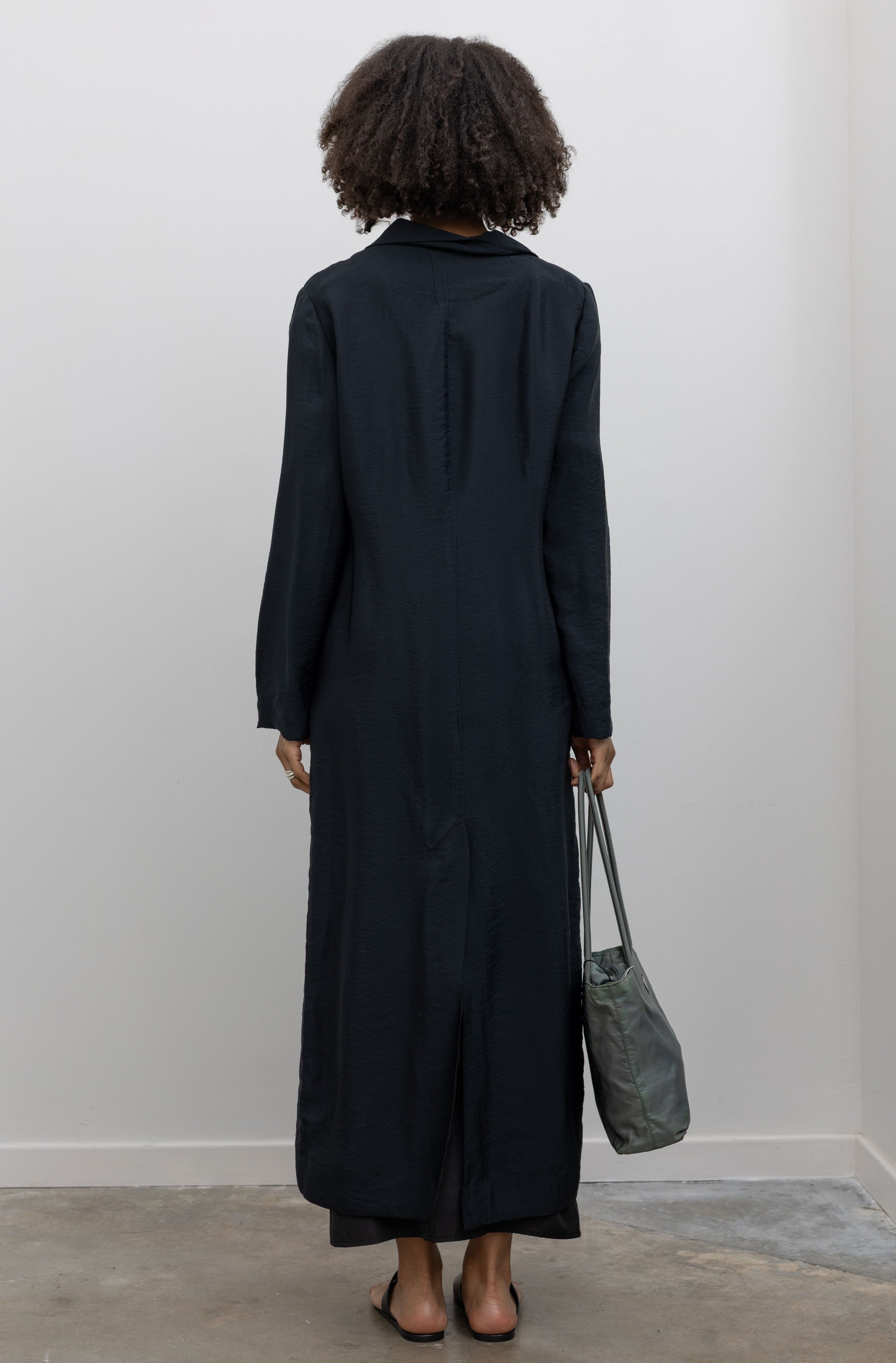 Full length front view of a woman in motion wearing contemporary spring and summer womenswear fashion in a elegant Nomia Black Long Unlined Relaxed Fit Duster Jacket with 6 front buttons and 2 patch pockets on front hip at an Alas Eius store. Made in New York from a beautiful Japanese Acetate blend with a washed vintage finish.