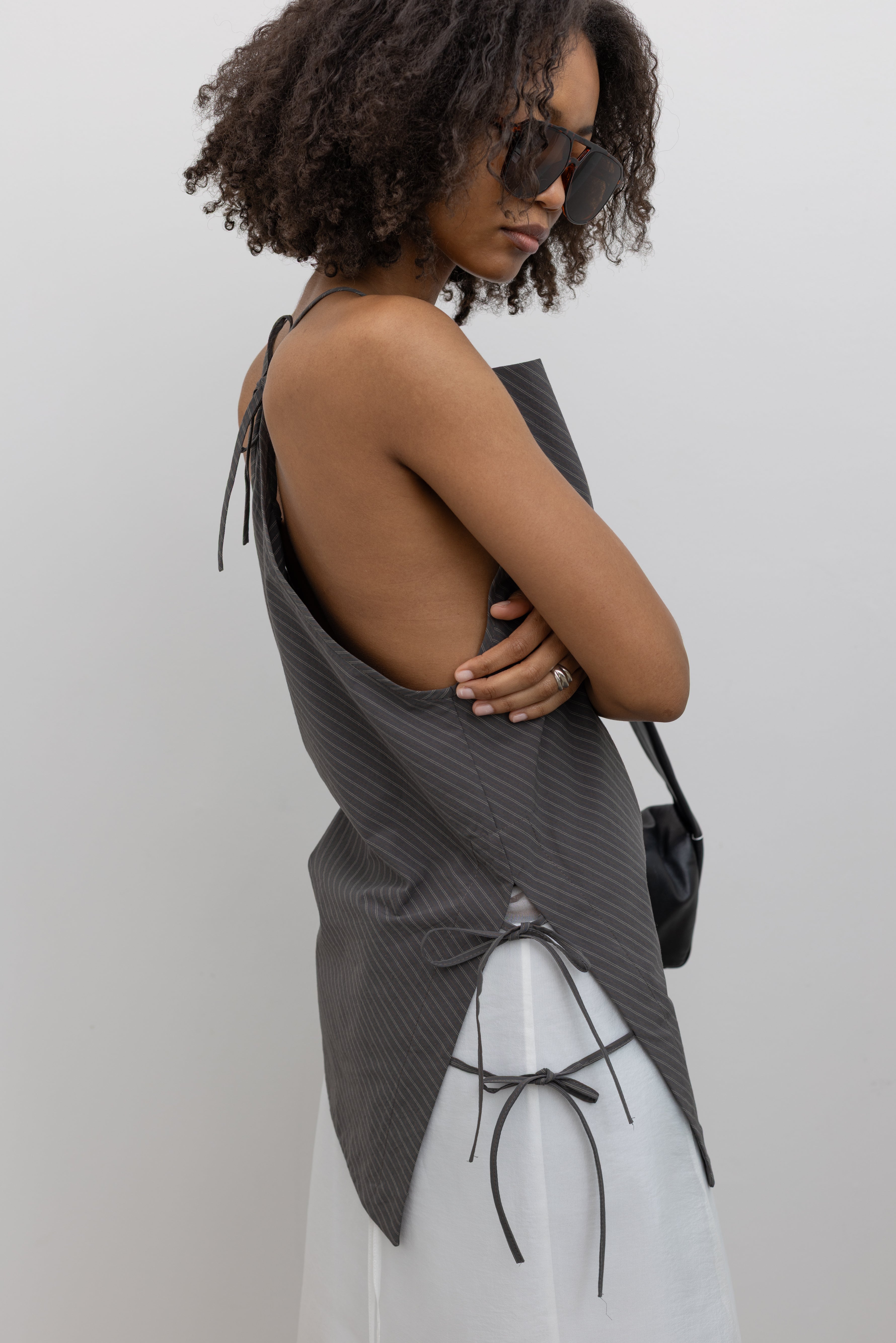 Close-up side view of a woman standing wearing modern minimalist spring and summer fashion in a chic Nomia coffee stripe side tie halter top featuring an asymmetrical hem and side ties on right hip. Slips over the head, with a keyhole at center back and adjustable spaghetti strap ties at an Alas Eius store. Made in New york City in a lightweight striped fabric from Japan.