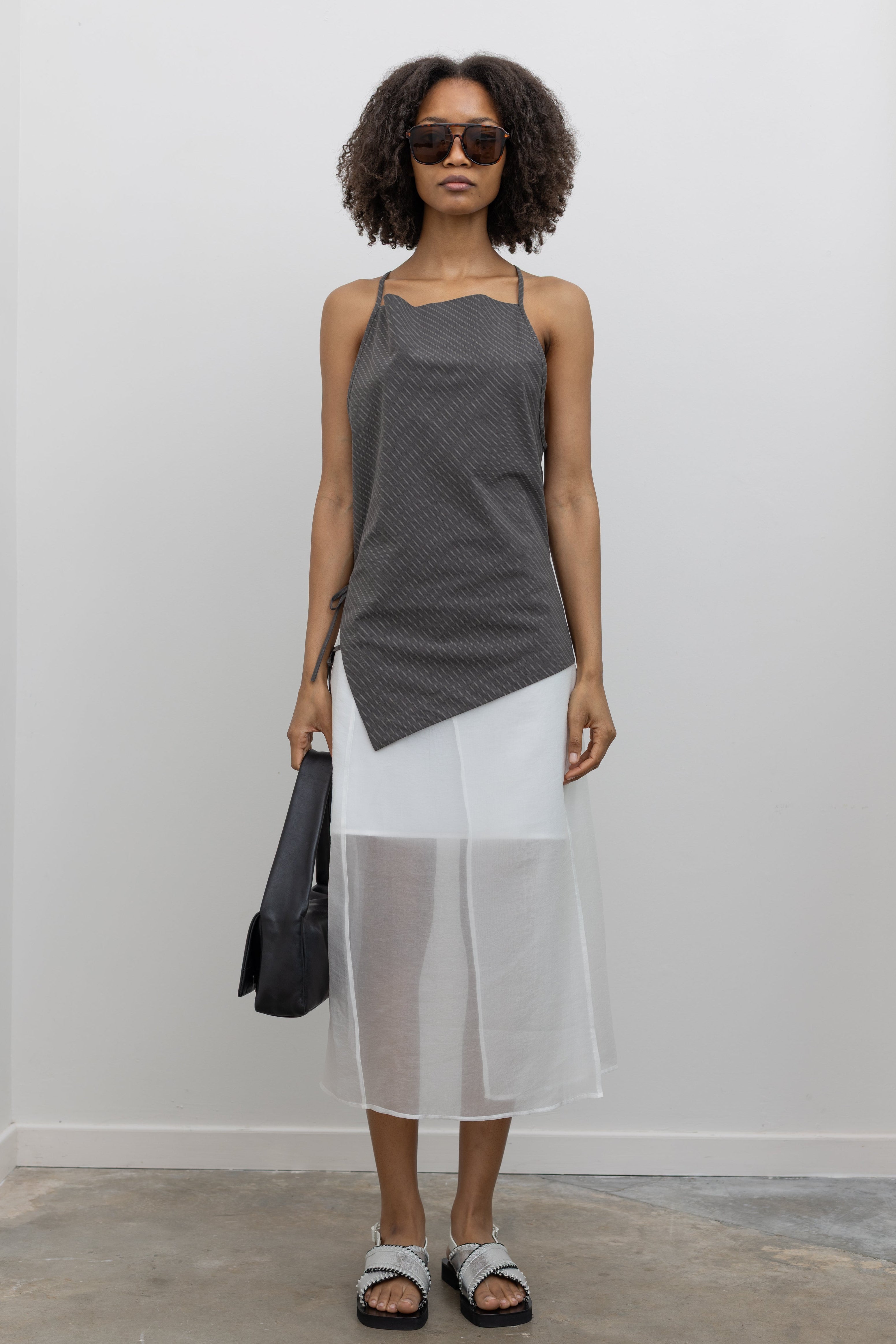 Full length front view of a woman standing wearing modern minimalist spring and summer fashion in a chic Nomia coffee stripe side tie halter top featuring an asymmetrical hem and side ties on right hip. Slips over the head, with a keyhole at center back and adjustable spaghetti strap ties at an Alas Eius store. Made in New york City in a lightweight striped fabric from Japan.