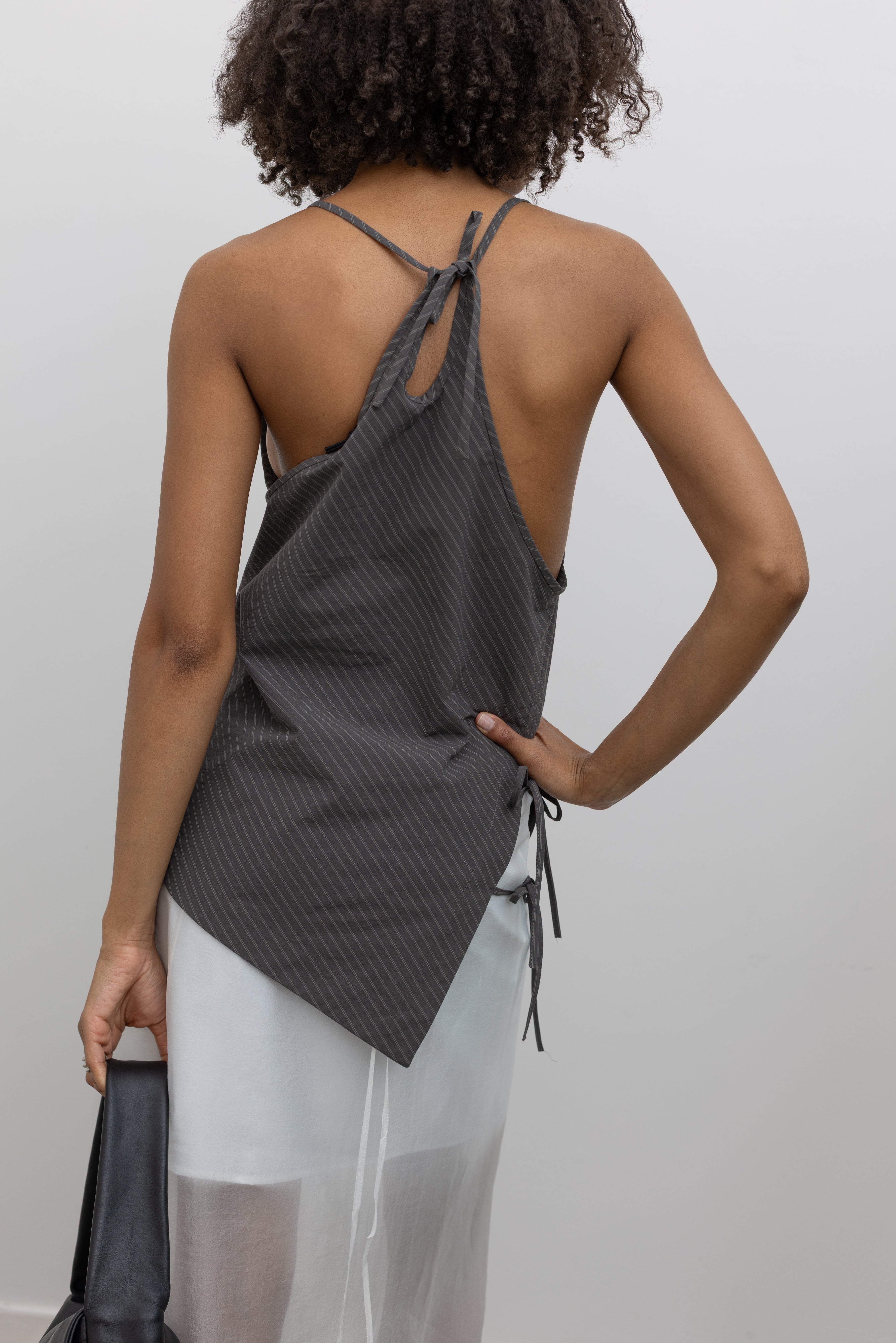 Close-up back view of a woman standing wearing modern minimalist spring and summer fashion in a chic Nomia coffee stripe side tie halter top featuring an asymmetrical hem and side ties on right hip. Slips over the head, with a keyhole at center back and adjustable spaghetti strap ties at an Alas Eius store. Made in New york City in a lightweight striped fabric from Japan.