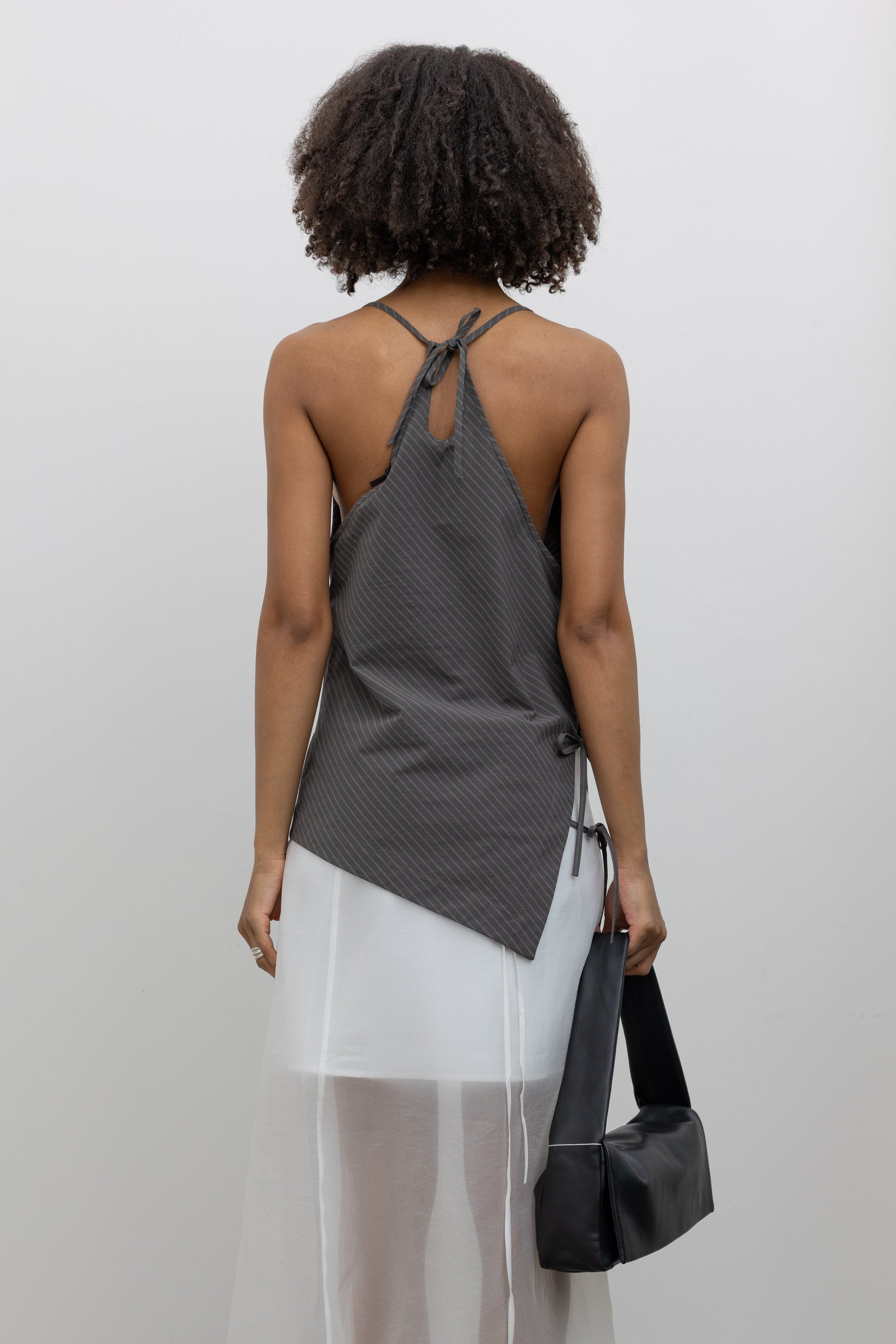 Close-up back view of a woman standing wearing modern minimalist spring and summer fashion in a chic Nomia coffee stripe side tie halter top featuring an asymmetrical hem and side ties on right hip. Slips over the head, with a keyhole at center back and adjustable spaghetti strap ties at an Alas Eius store. Made in New york City in a lightweight striped fabric from Japan.