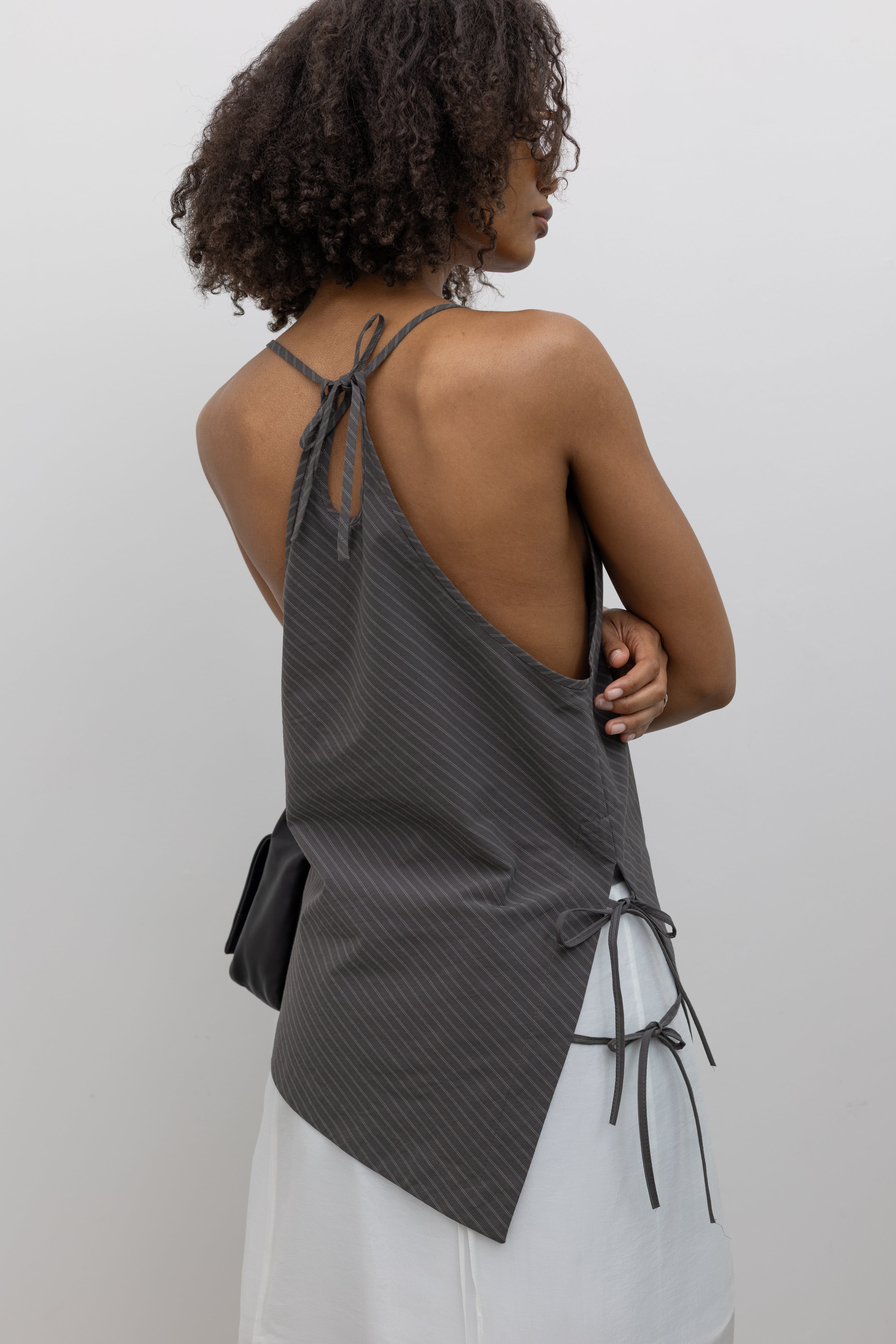 Close-up back view of a woman standing wearing contemporary spring and summer fashion in a chic Nomia coffee stripe side tie halter top featuring an asymmetrical hem and side ties on right hip. Slips over the head, with a keyhole at center back and adjustable spaghetti strap ties at an Alas Eius store. Made in New york City in a lightweight striped fabric from Japan.