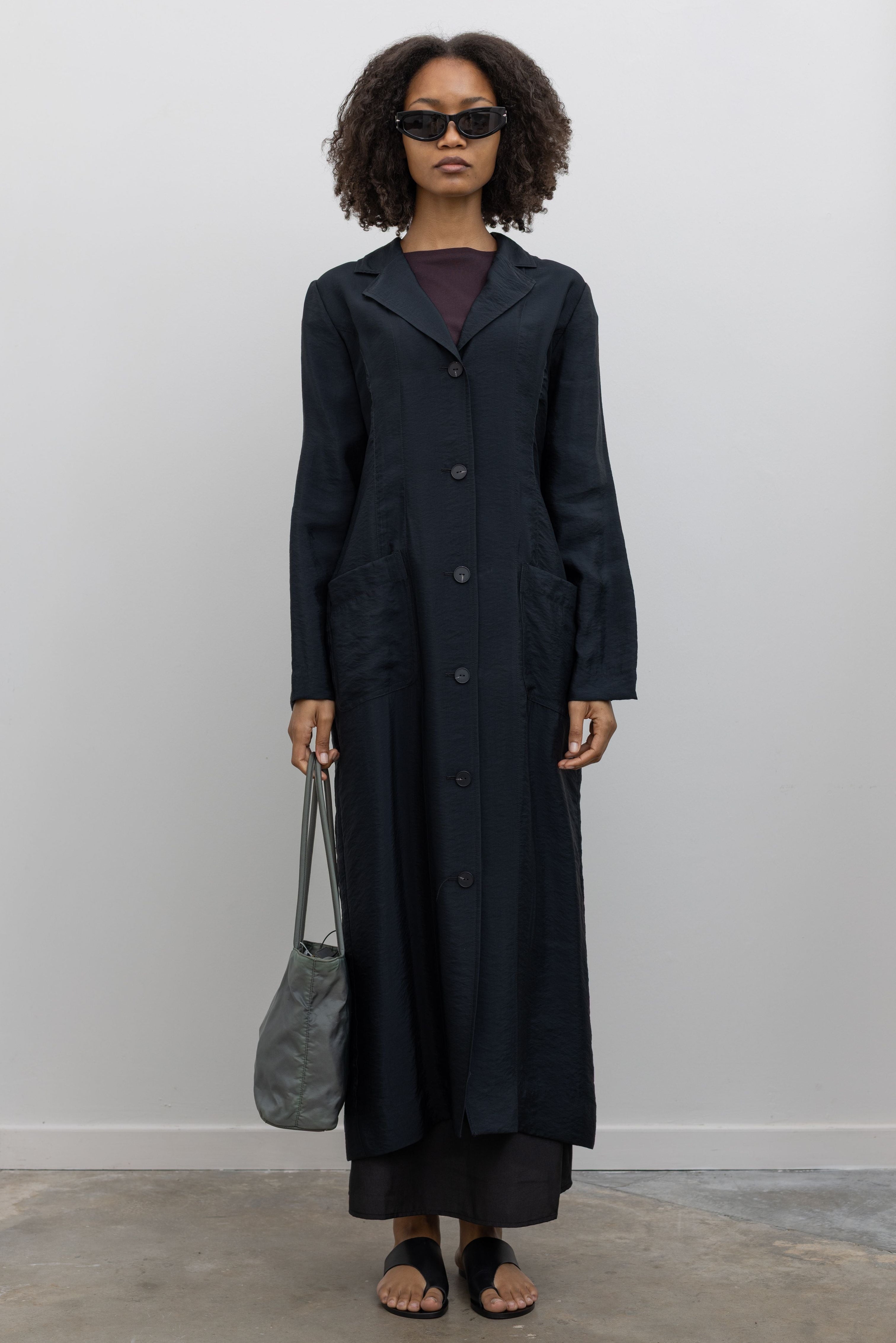 Full length front view of a woman in motion wearing modern minimalist spring and summer womenswear fashion in a elegant Nomia Black Long Unlined Relaxed Fit Duster Jacket with 6 front buttons and 2 patch pockets on front hip at an Alas Eius store. Made in New York from a beautiful Japanese Acetate blend with a washed vintage finish.
