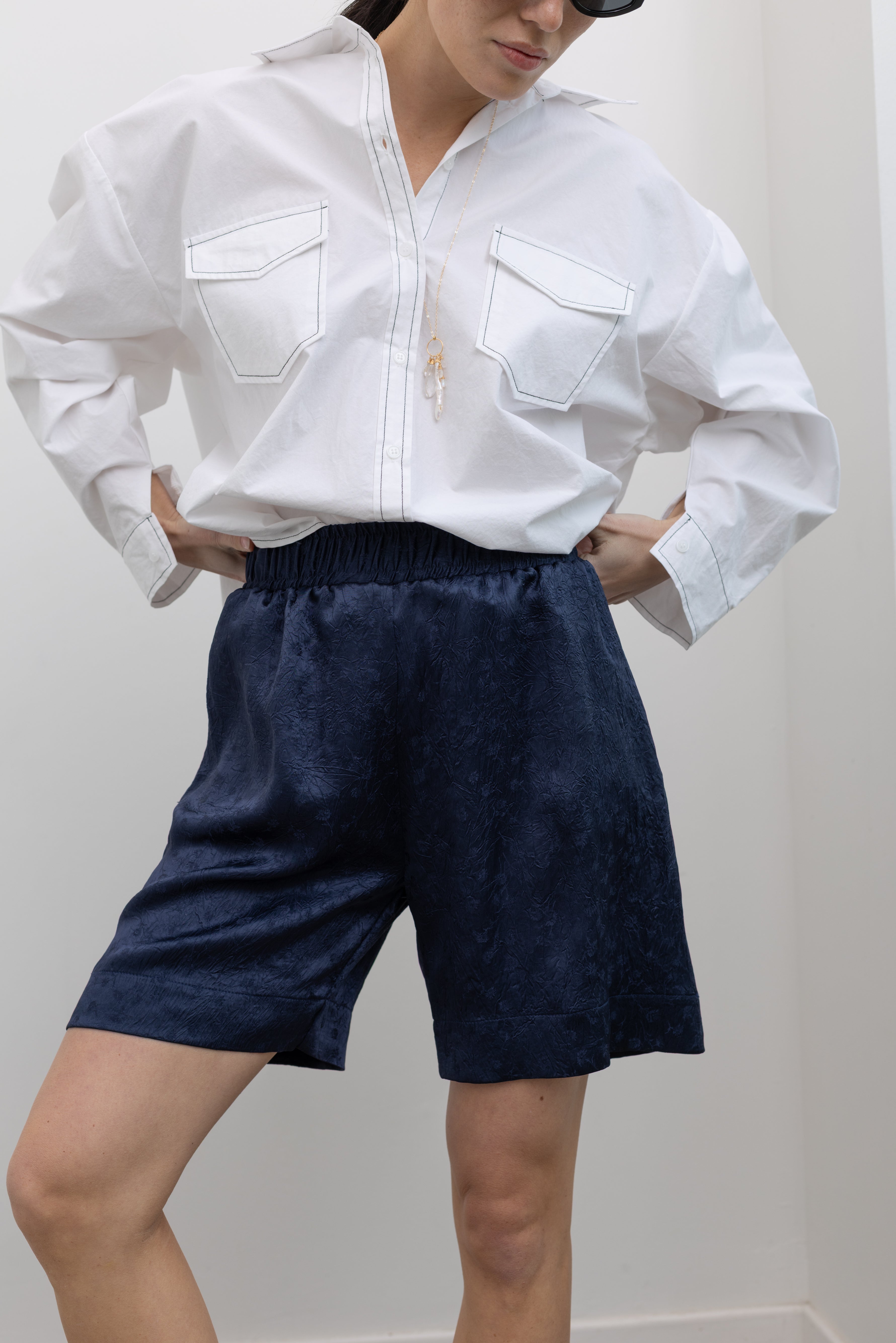 Close-up front view of a woman wearing elevated spring fashion in an Alas Eius label classic white button down shirt with contrasting black stitching paired with the Nomia deep ink blue floral jacquard boxer shorts featuring an elastic waistband and side seam pockets at Alas Eius. 