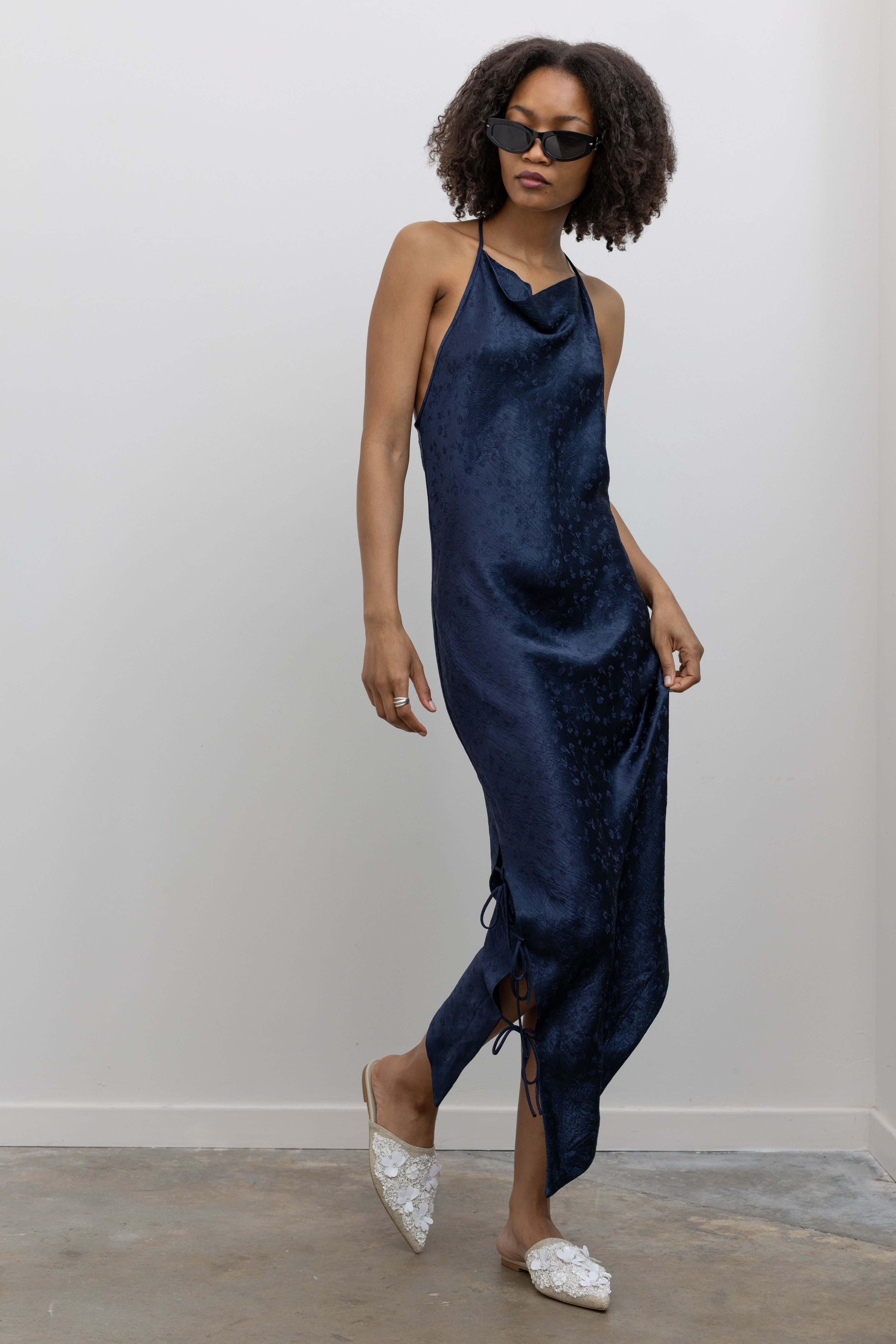 Full length 3/4 view of a woman standing wearing contemporary spring and summer fashion in a Nomia ink floral jacquard side tie bias cut dress featuring an adjustable cowl neckline, and an asymmetrical hem at an Alas Eius store. Made in New York City, USA.