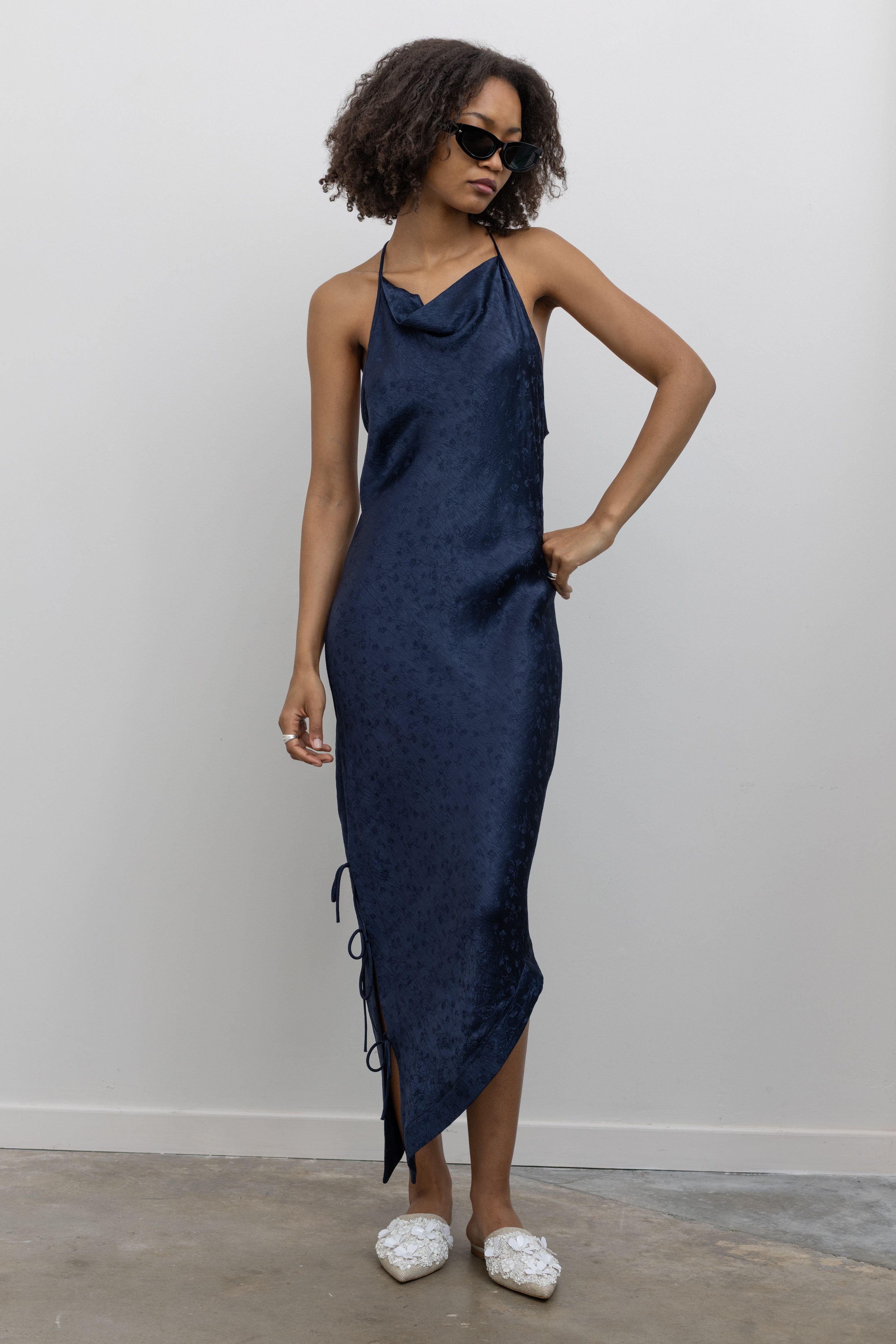 Full length front view of a woman standing wearing contemporary spring and summer fashion in a Nomia ink floral jacquard side tie bias cut dress featuring an adjustable cowl neckline, and an asymmetrical hem at an Alas Eius store. Made in New York City, USA.