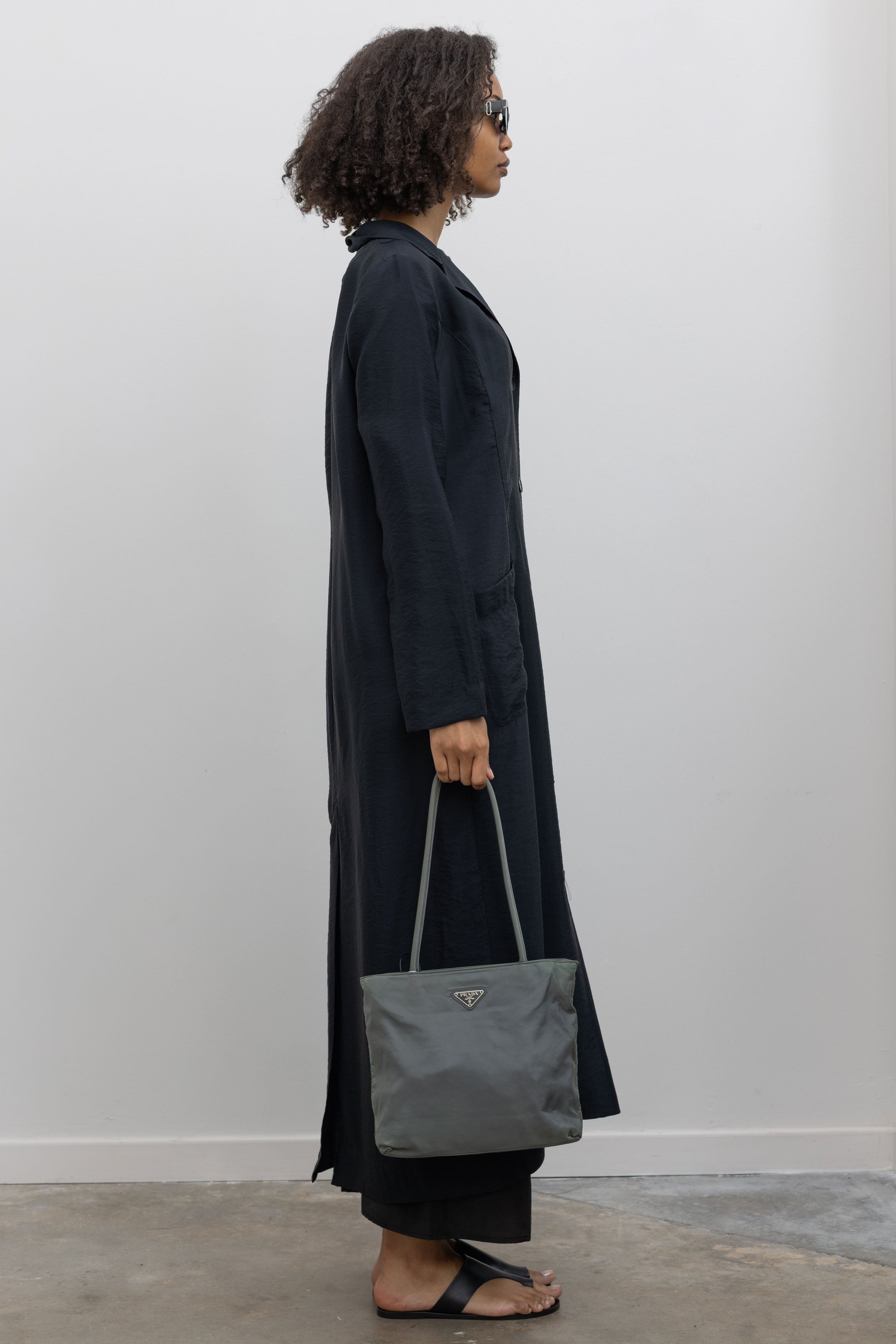 Full length side view of a woman in motion wearing modern minimalist spring and summer womenswear fashion in a elegant Nomia Black Long Unlined Relaxed Fit Duster Jacket with 6 front buttons and 2 patch pockets on front hip at an Alas Eius store. Made in New York from a beautiful Japanese Acetate blend with a washed vintage finish.