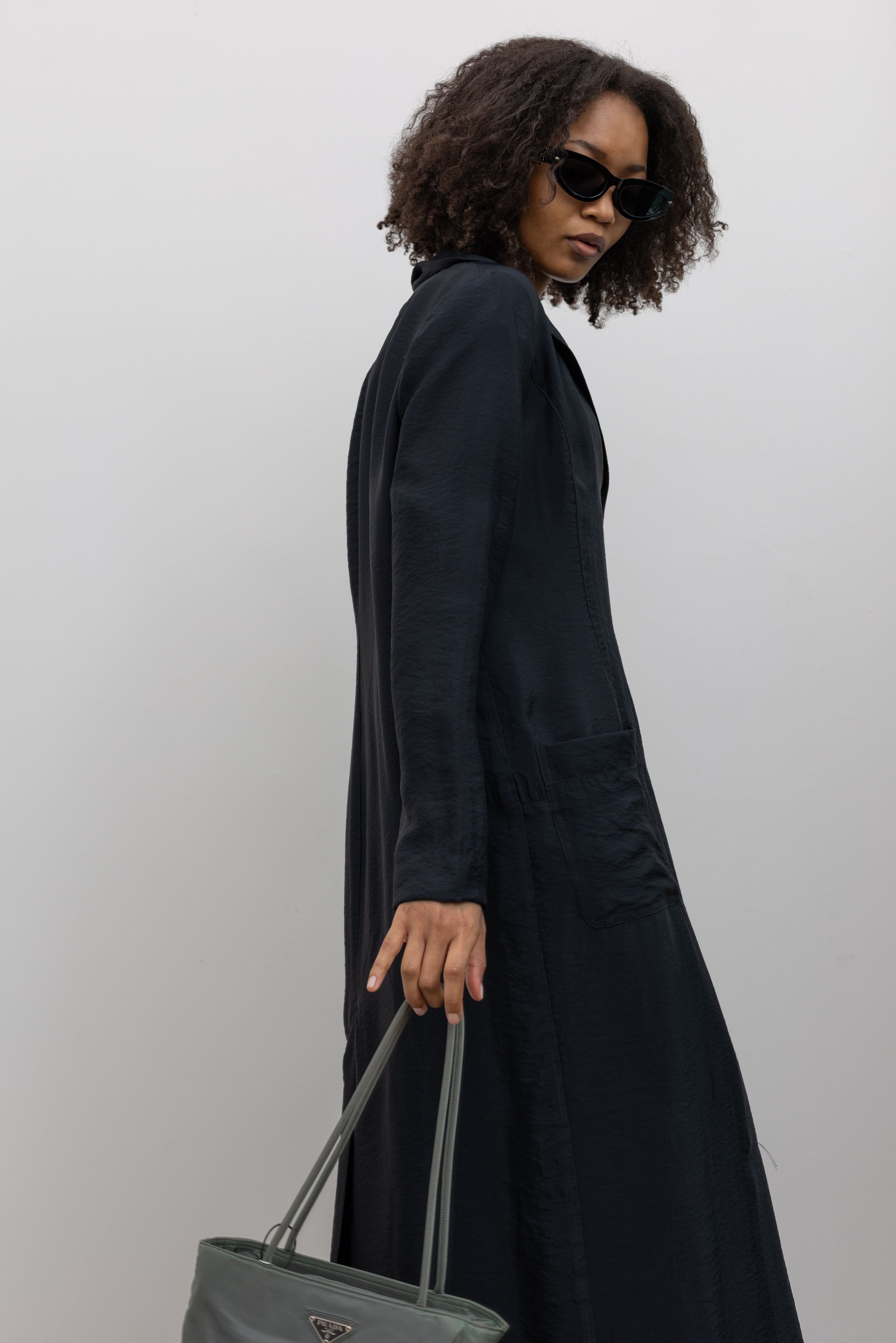 3/4 view of a woman in motion wearing modern and modest spring and summer womenswear fashion in a elegant Nomia Black Long Unlined Relaxed Fit Duster Jacket with 6 front buttons and 2 patch pockets on front hip at an Alas Eius store. Made in New York from a beautiful Japanese Acetate blend with a washed vintage finish.