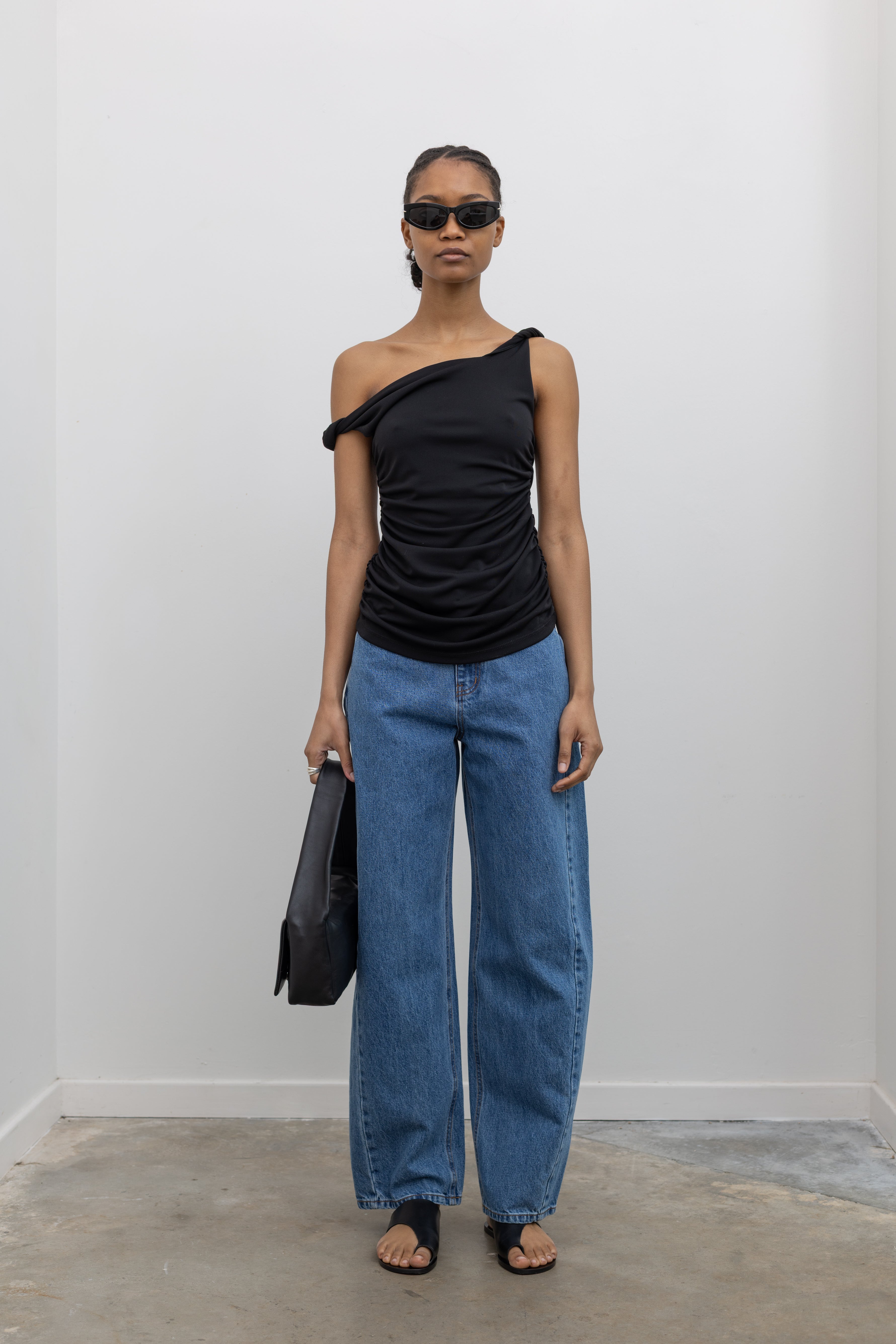 Full front view of a woman standing wearing Spring fashion in light wash denim jeans paired with a flattering Nomia black twisted off the shoulder straps and shirring detail on side seams at Alas Eius.