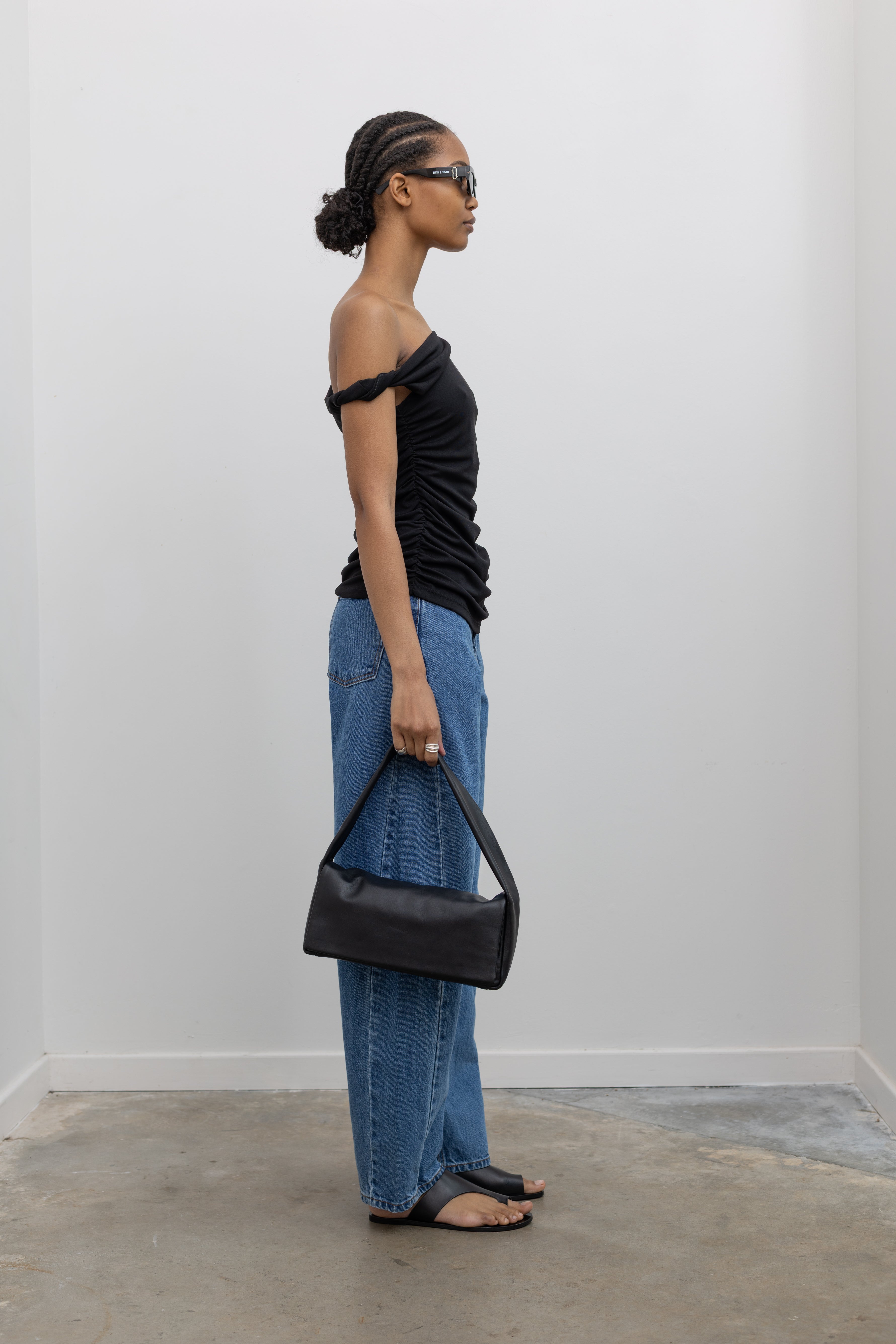 Full length side view of a woman wearing Spring fashion in light wash denim jeans, a St. Agni leather bag, and ATP black leather cutout sandals paired with a flattering Nomia black twisted off the shoulder straps and shirring detail on side seams at Alas Eius.