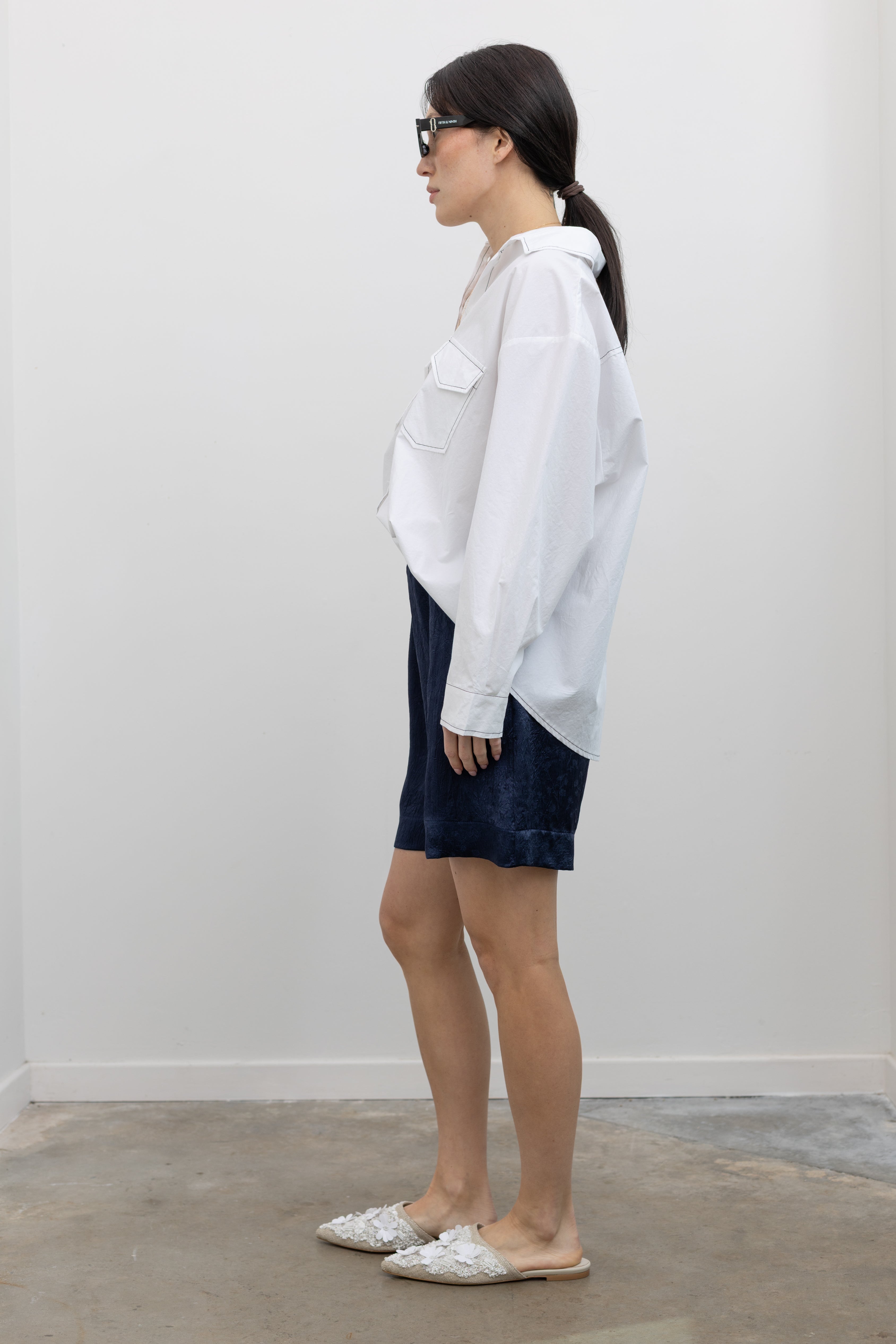 Full length side view of a woman wearing elevated spring fashion in an Alas Eius label classic white button down shirt with contrasting black stitching paired with the Nomia deep ink blue floral jacquard boxer shorts featuring an elastic waistband and side seam pockets at Alas Eius. 