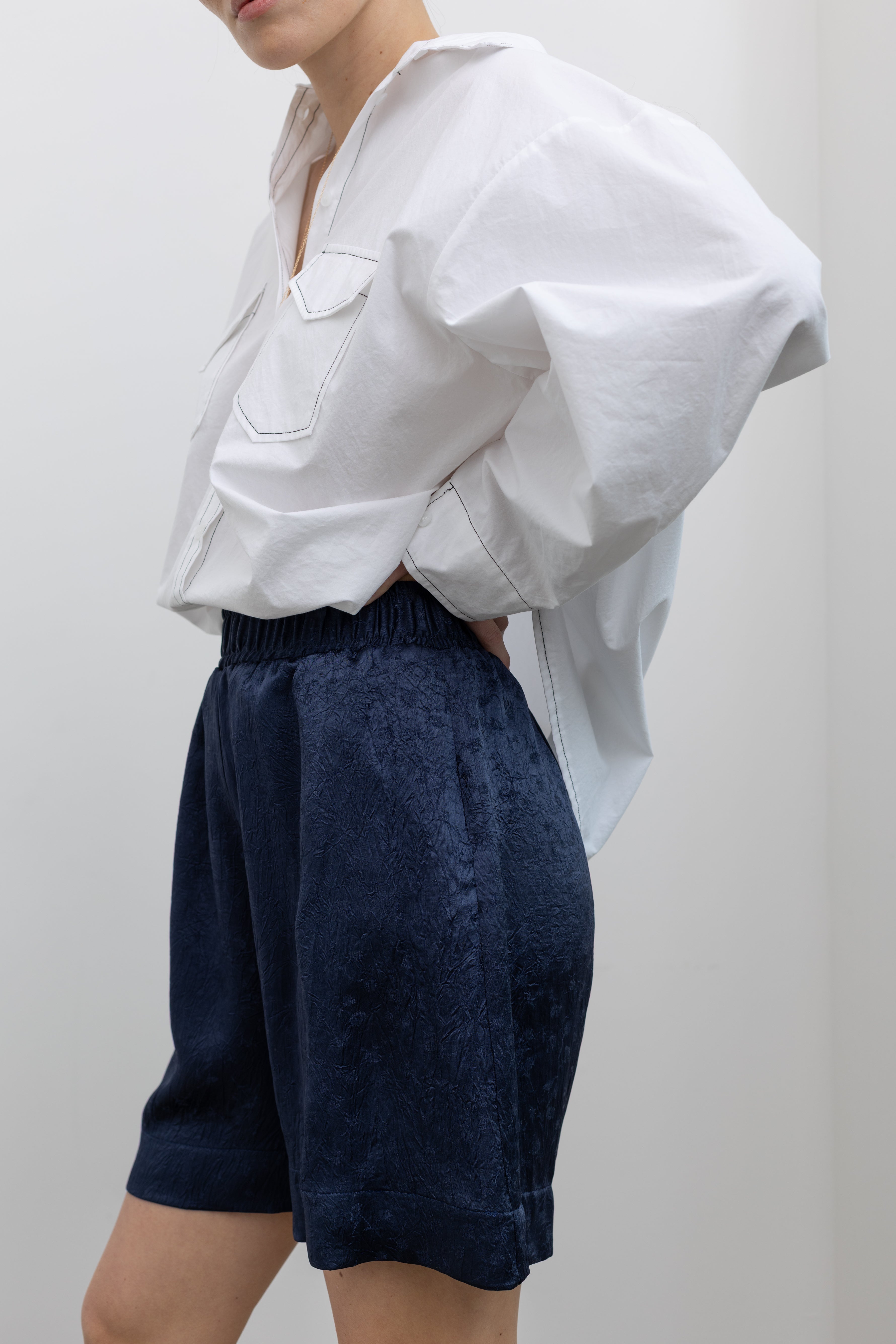 Close-up side view of a woman wearing elevated spring fashion in an Alas Eius label classic white button down shirt with contrasting black stitching paired with the Nomia deep ink blue floral jacquard boxer shorts featuring an elastic waistband and side seam pockets at Alas Eius. 