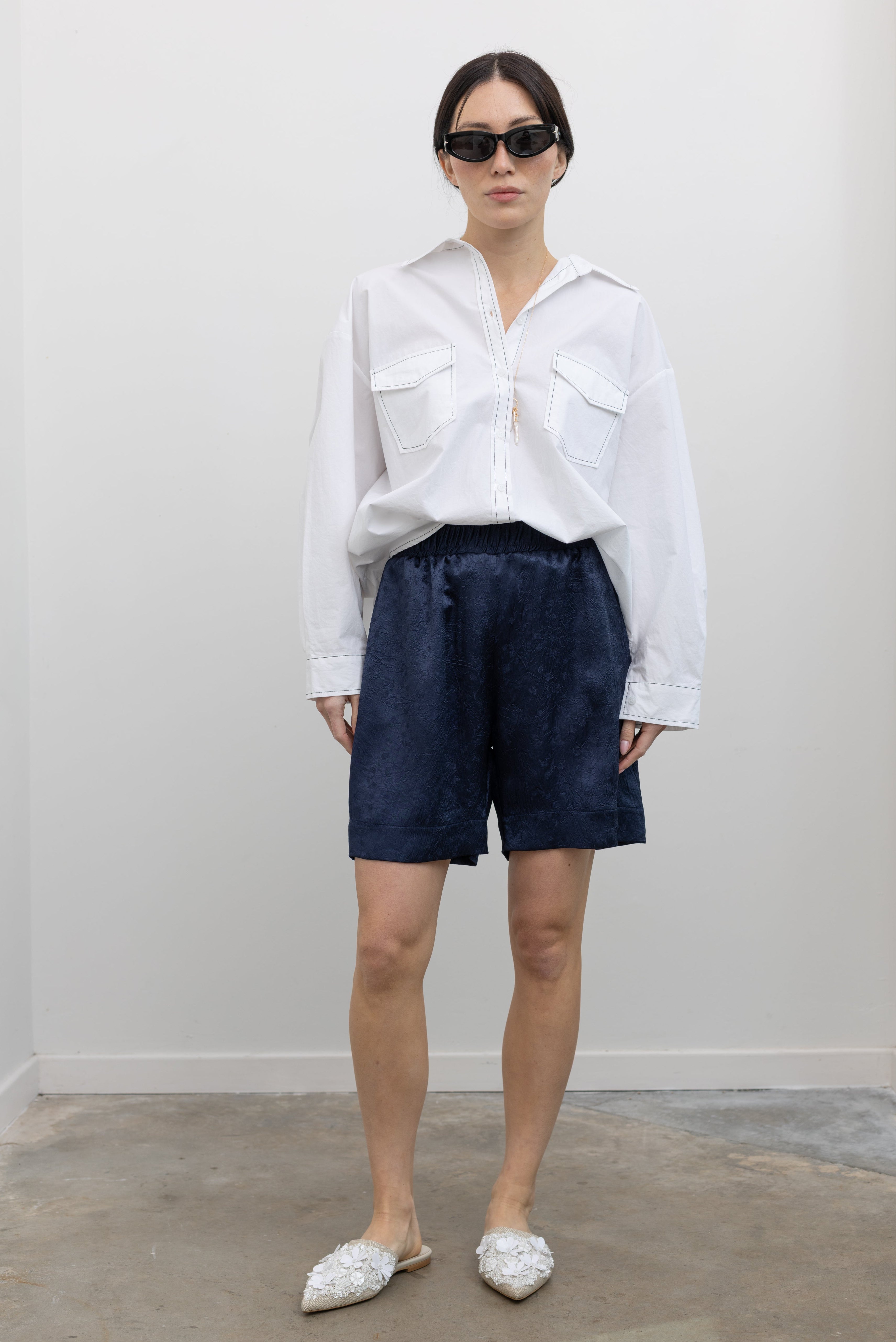 Full length front view of a woman wearing elevated spring fashion in an Alas Eius label classic white button down shirt with contrasting black stitching paired with the Nomia deep ink blue floral jacquard boxer shorts featuring an elastic waistband and side seam pockets at Alas Eius. 