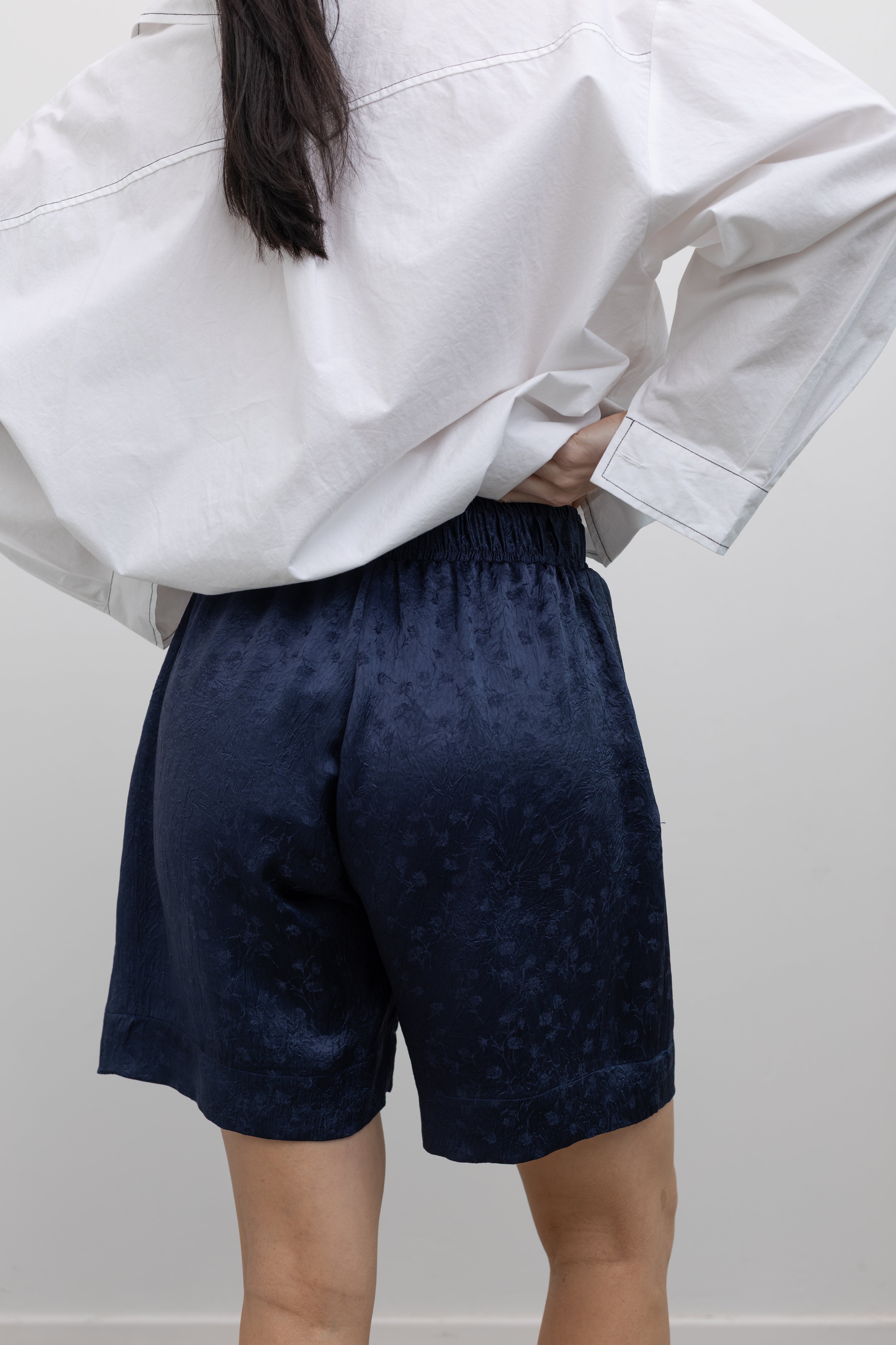 Close-up back view of a woman wearing elevated spring fashion in an Alas Eius label classic white button down shirt with contrasting black stitching paired with the Nomia deep ink blue floral jacquard boxer shorts featuring an elastic waistband and side seam pockets at Alas Eius. 