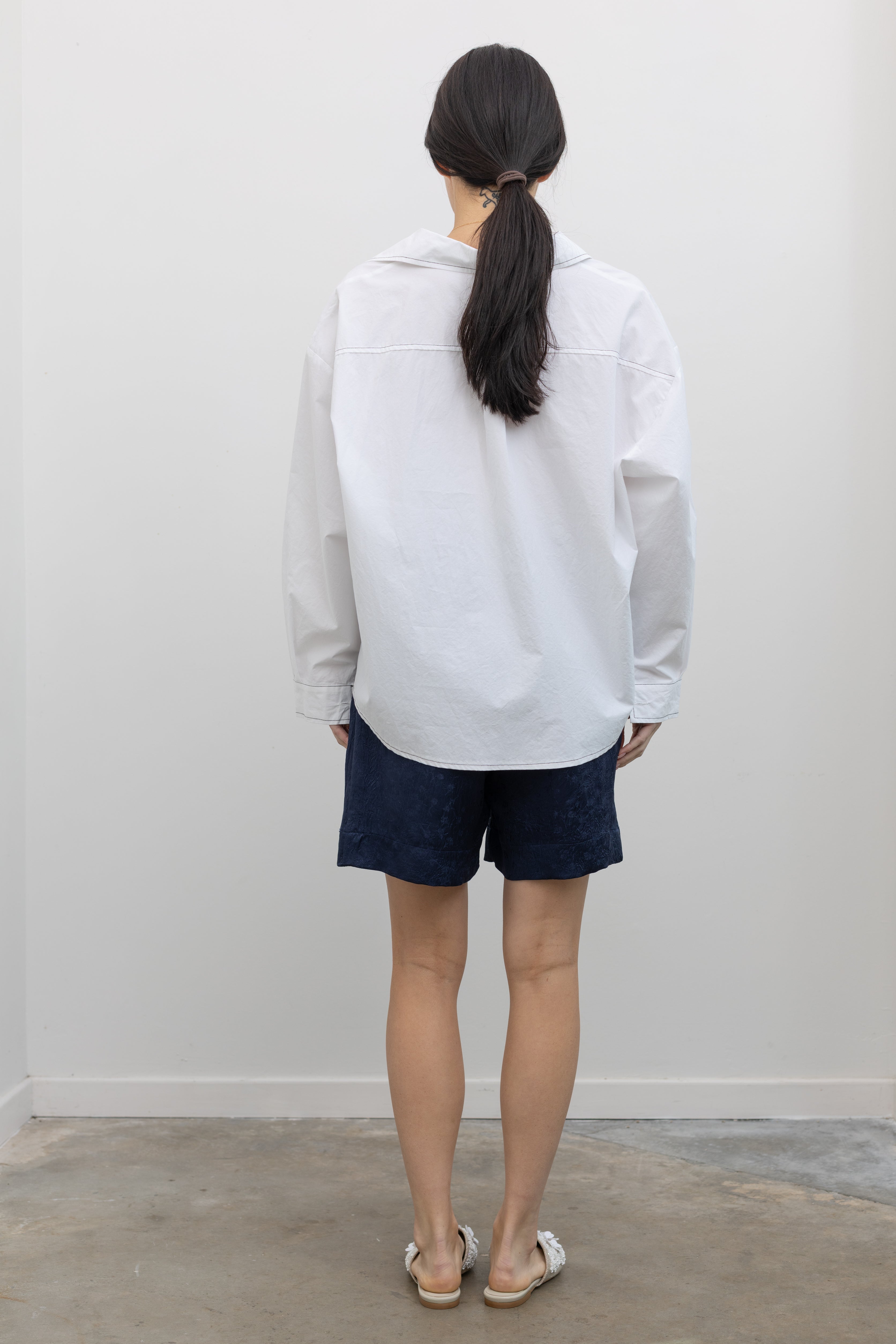 Full length back view of a woman wearing elevated spring fashion in an Alas Eius label classic white button down shirt with contrasting black stitching paired with the Nomia deep ink blue floral jacquard boxer shorts featuring an elastic waistband and side seam pockets at Alas Eius. 
