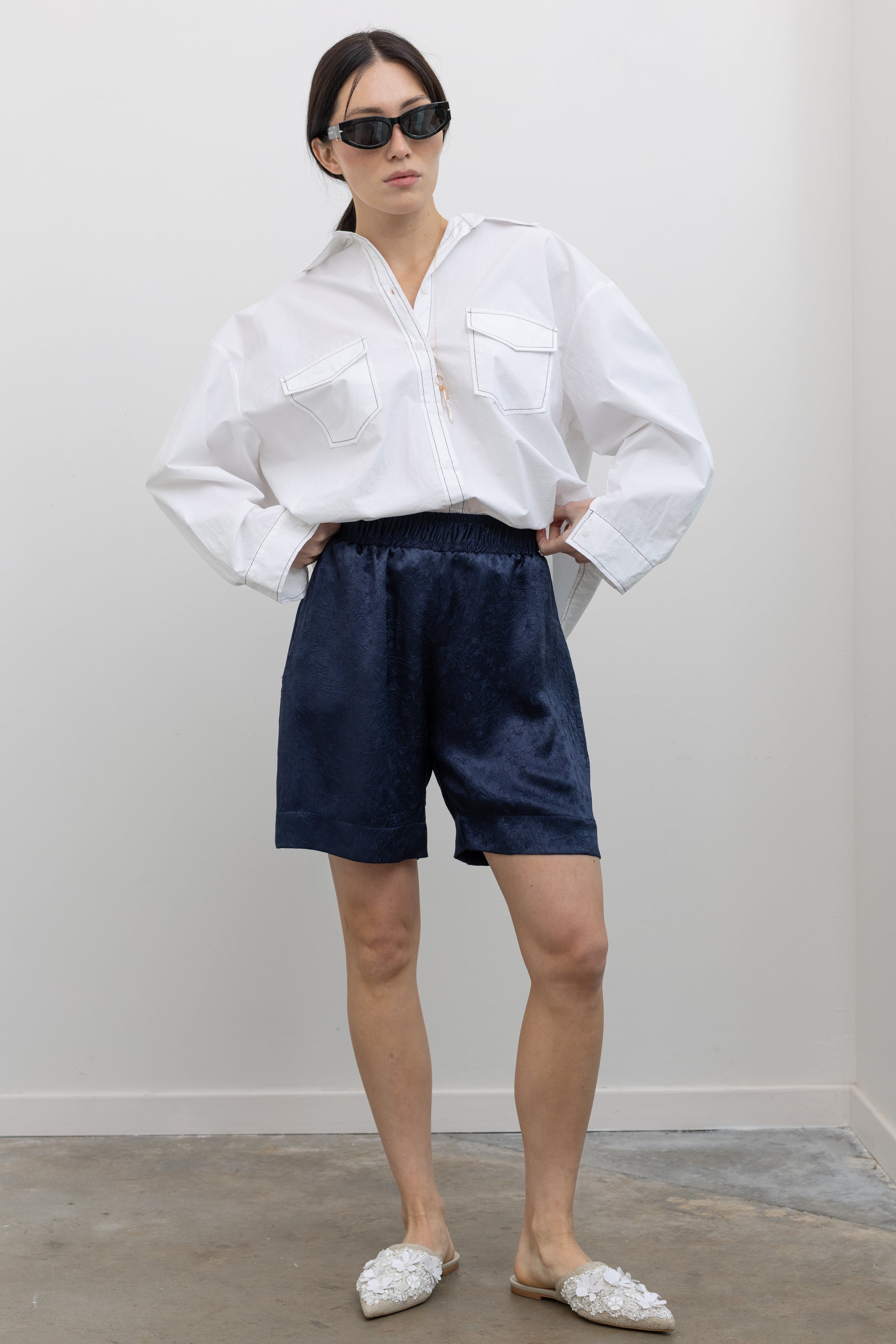Full length front view of a woman wearing elevated spring fashion in an Alas Eius label classic white button down shirt with contrasting black stitching paired with the Nomia deep ink blue floral jacquard boxer shorts featuring an elastic waistband and side seam pockets at Alas Eius. 
