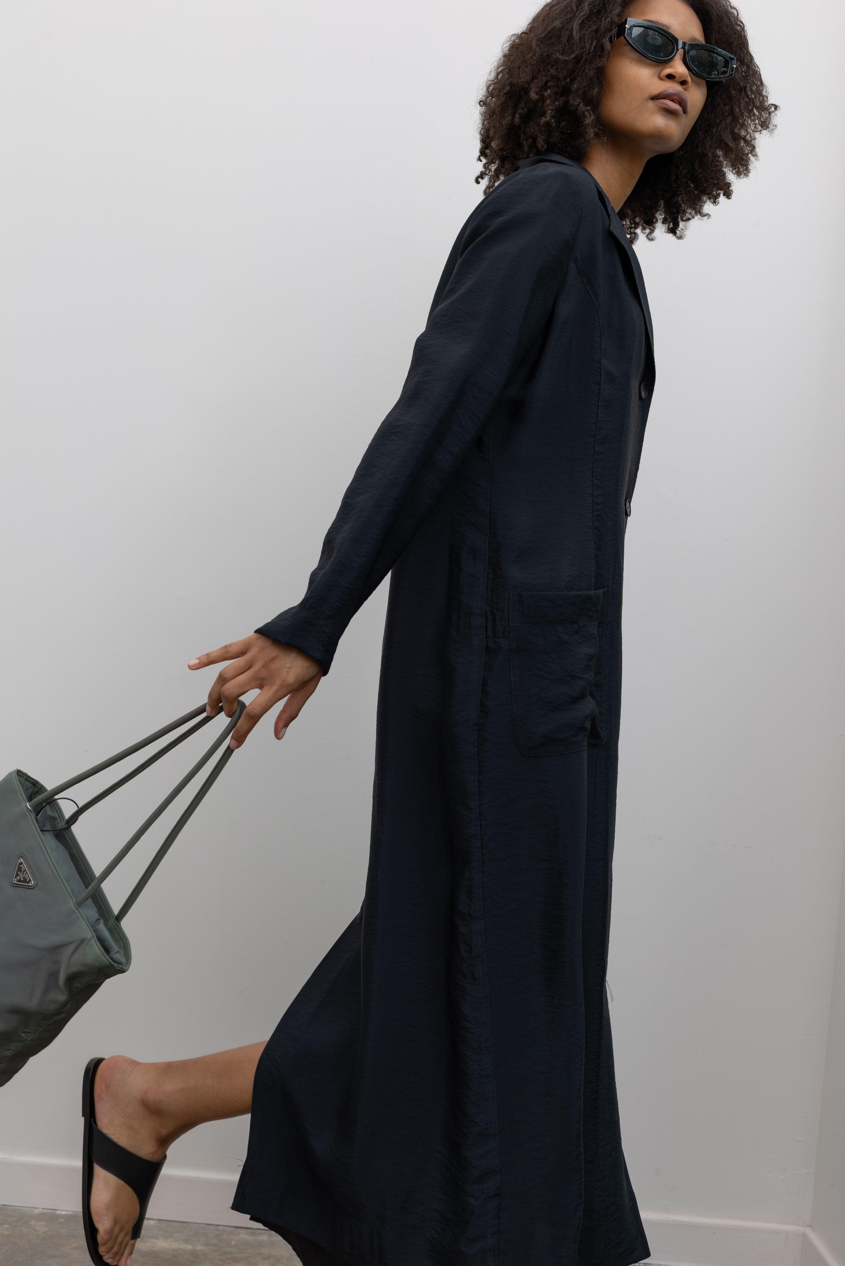 Side view of a woman in motion wearing modern minimalist spring and summer womenswear fashion in a elegant Nomia Black Long Unlined Relaxed Fit Duster Jacket with 6 front buttons and 2 patch pockets on front hip at an Alas Eius store. Made in New York from a beautiful Japanese Acetate blend with a washed vintage finish.