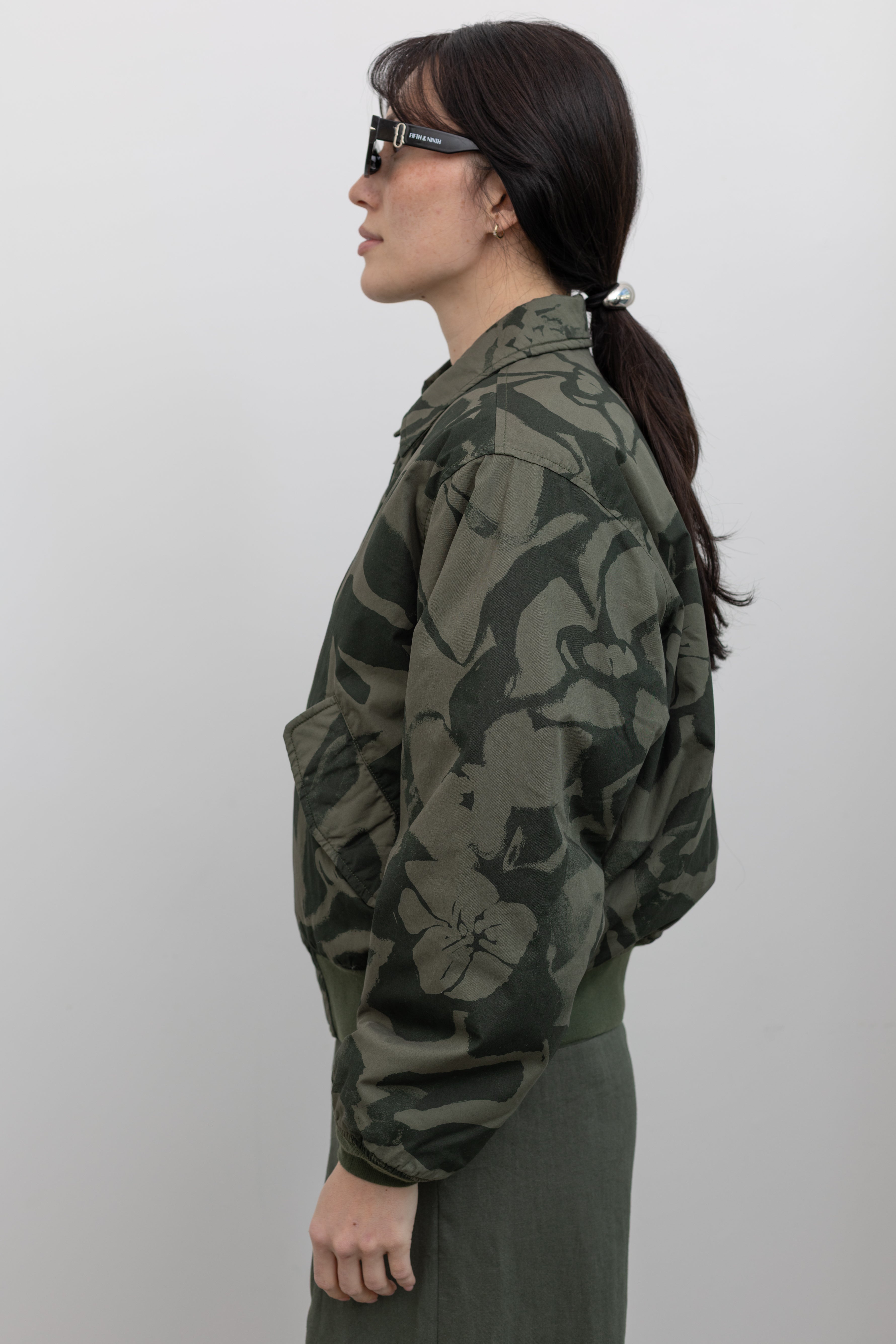 Close-up side view of a woman standing wearing spring fashion in a refined and functional Obey brand green floral print cotton bomber jacket with a silver front zipper and classic ribbed cuffs at an Alas Eius store. 