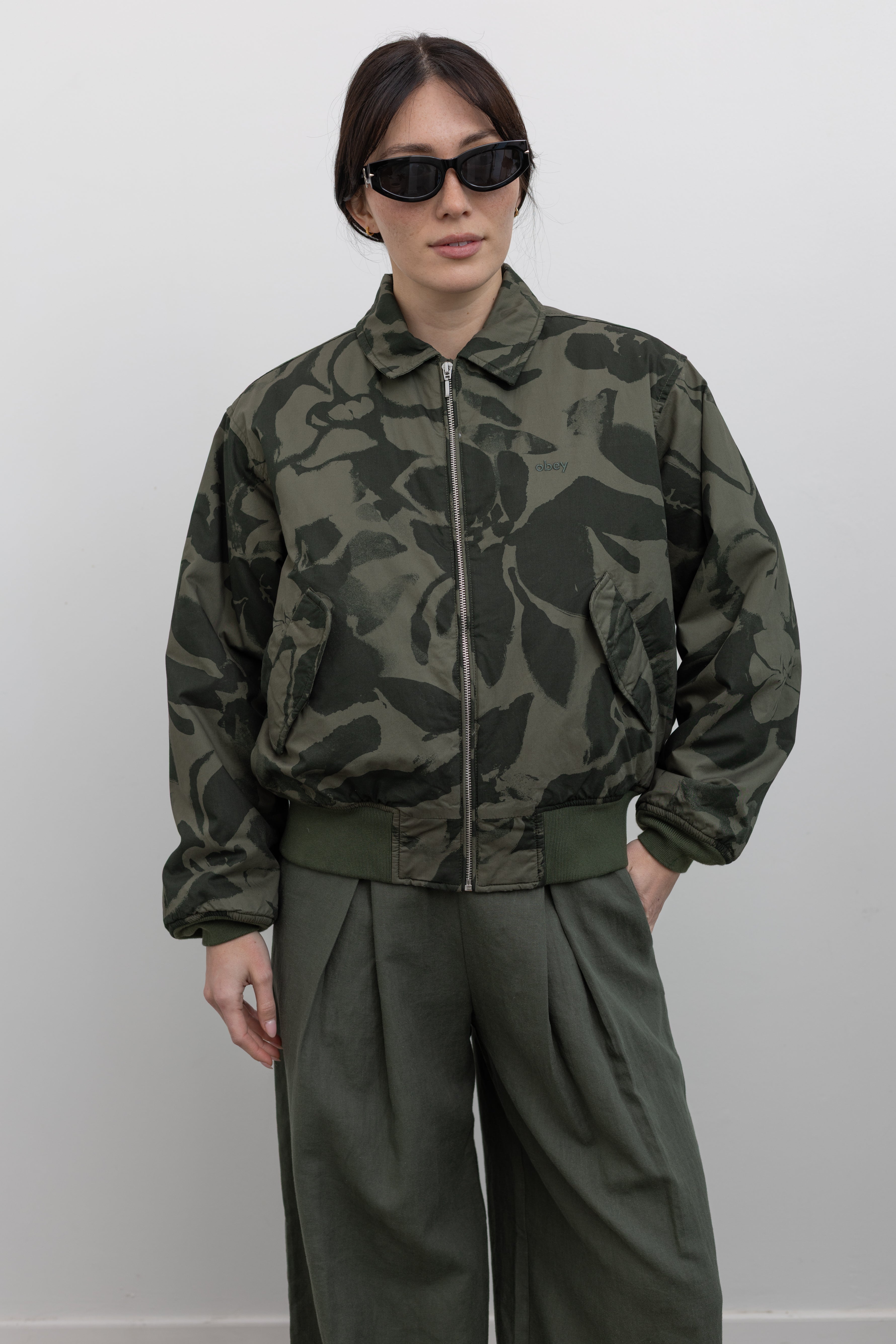 Woman standing wearing spring fashion in a chic and functional Obey brand green floral print cotton bomber jacket with a silver front zipper and classic ribbed cuffs at an Alas Eius store. 