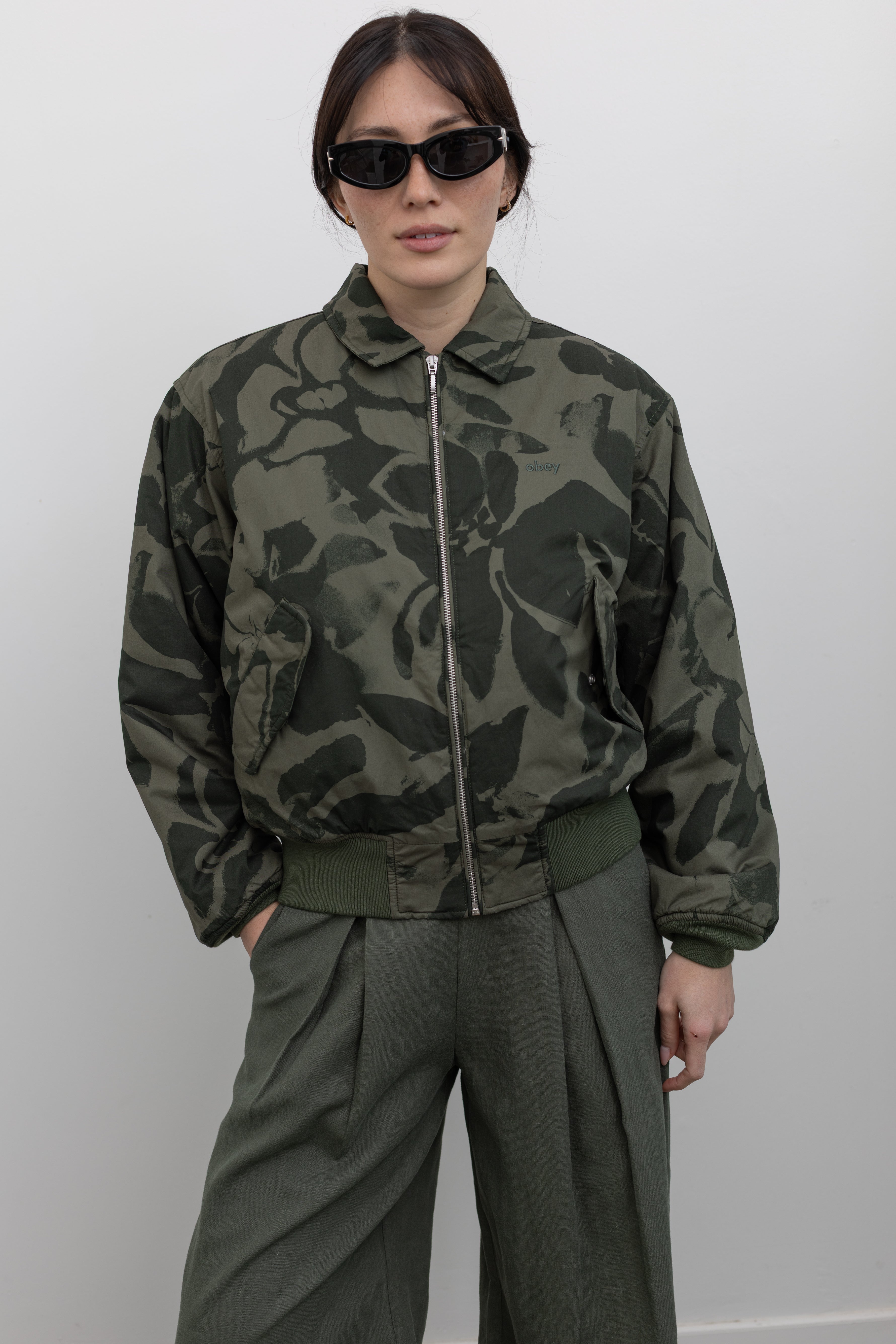 Close-up front view of a woman standing wearing spring fashion in a refined and functional Obey brand green floral print cotton bomber jacket with a silver front zipper and classic ribbed cuffs at an Alas Eius store. 