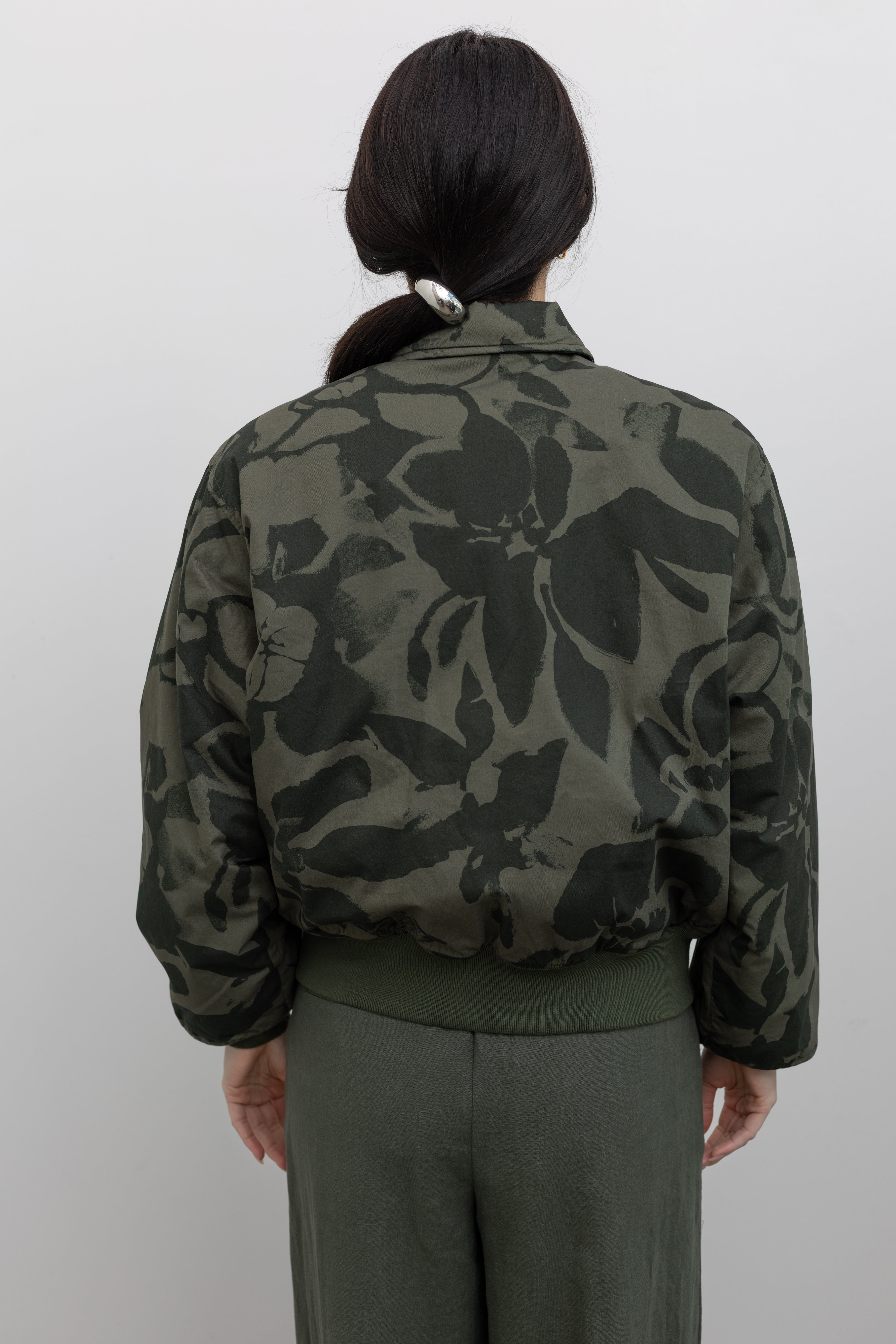 Close-up back view of a woman standing wearing spring fashion in a refined and functional Obey brand green floral print cotton bomber jacket with a silver front zipper and classic ribbed cuffs at an Alas Eius store. 