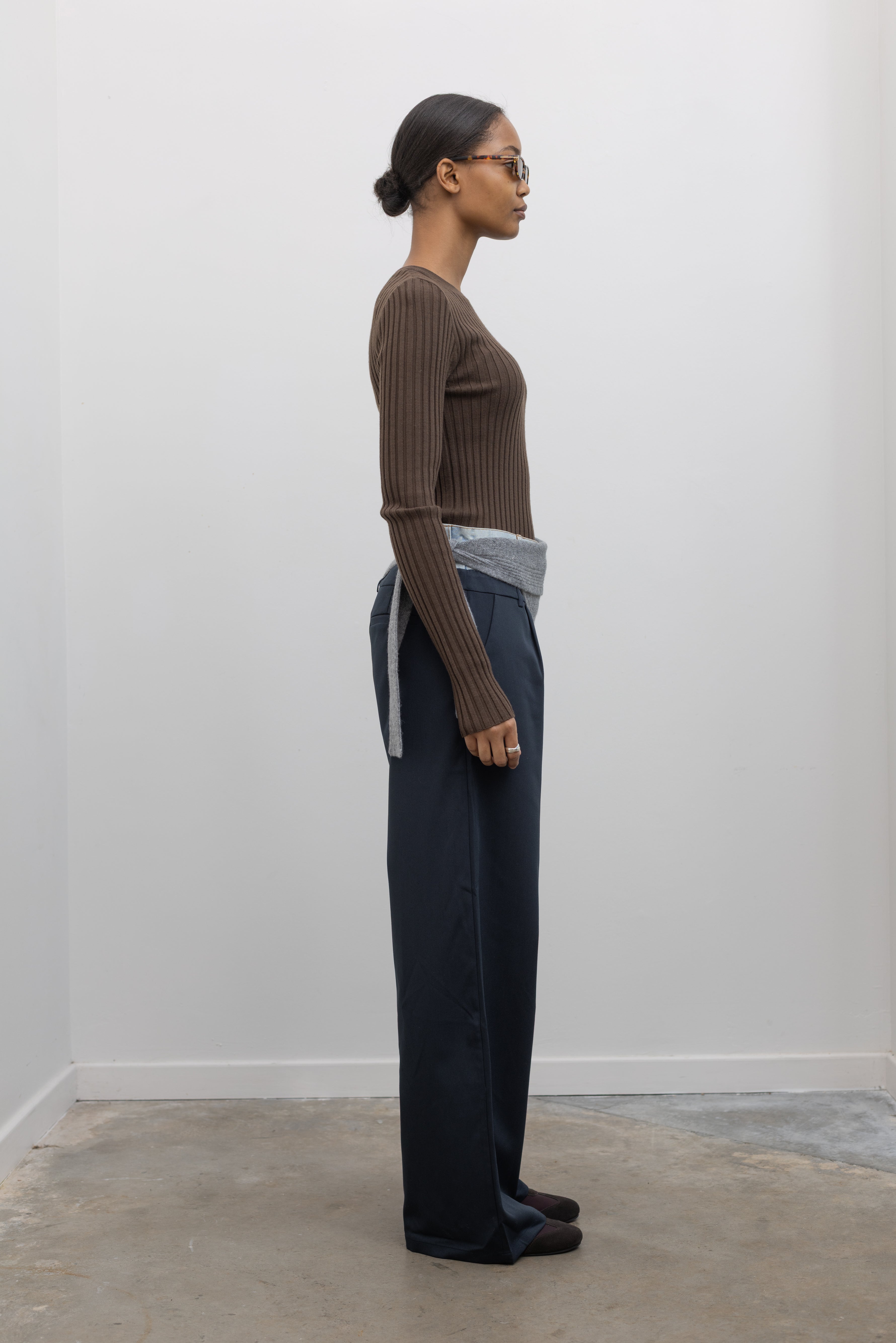 Full length side view of a woman standing wearing an elevated essential spring staple in a Paper Moon brand brown wool blend ribbed long sleeve fitted top with navy blue trousers and a gray bandana scarf around her waist at an Alas Eius store. 