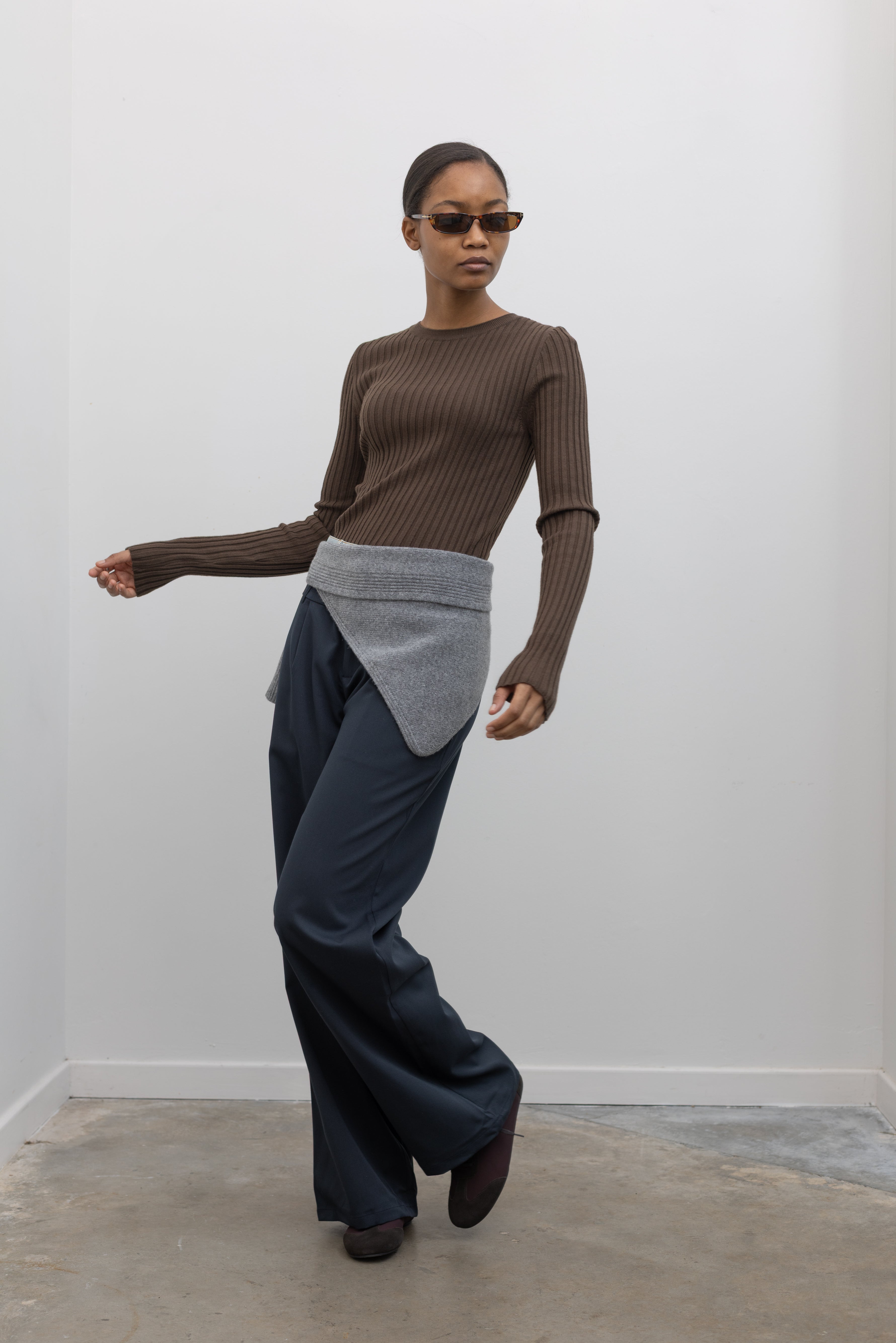 Full length side view of a woman in motion wearing an elevated essential spring staple in a Paper Moon brand brown wool blend ribbed long sleeve fitted top with navy blue trousers and a gray bandana scarf around her waist at an Alas Eius 