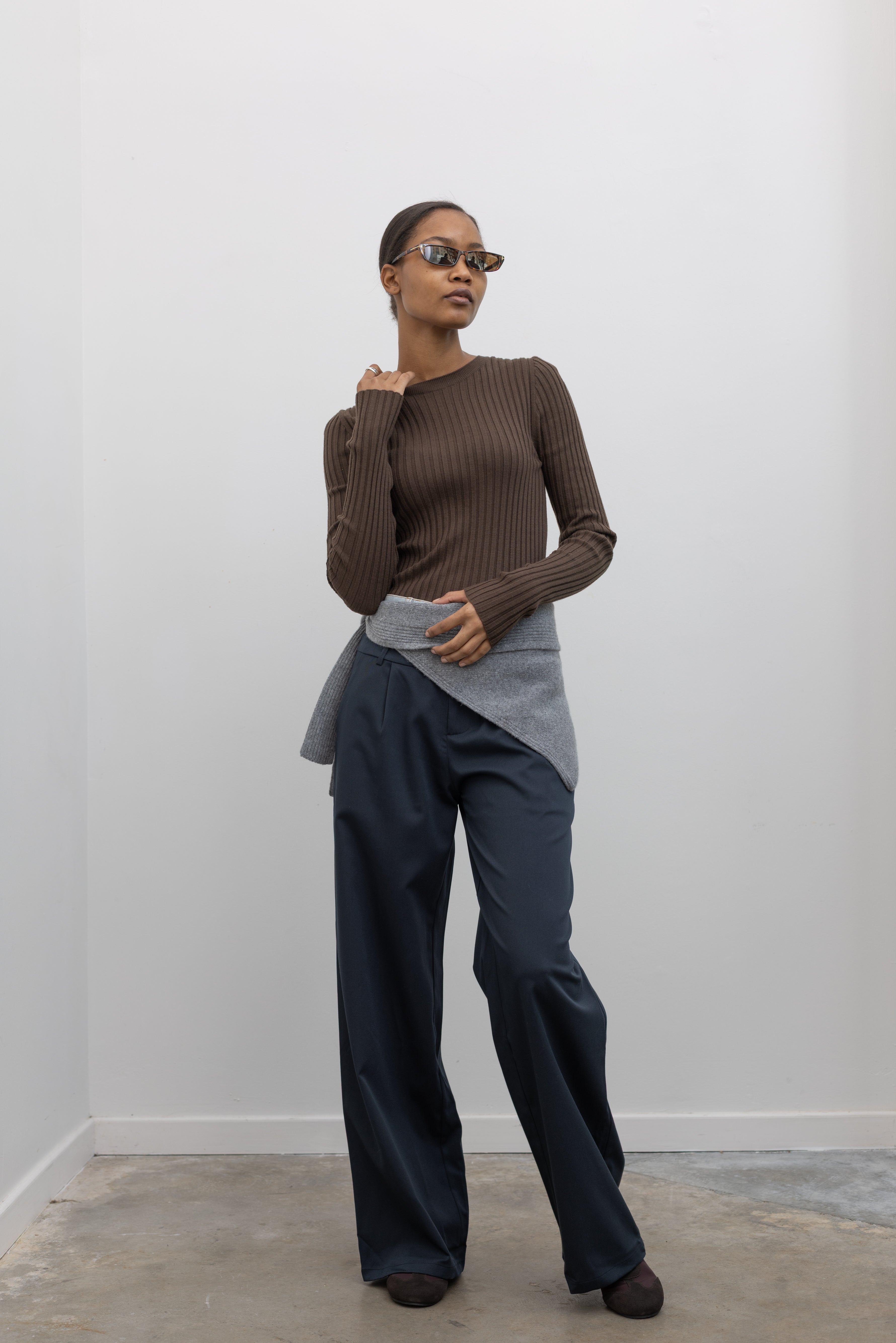 Full length front view of a woman standing wearing an elevated essential spring staple in a Paper Moon brand brown wool blend ribbed long sleeve fitted top with navy blue trousers and a gray bandana scarf around her waist at an Alas Eius 