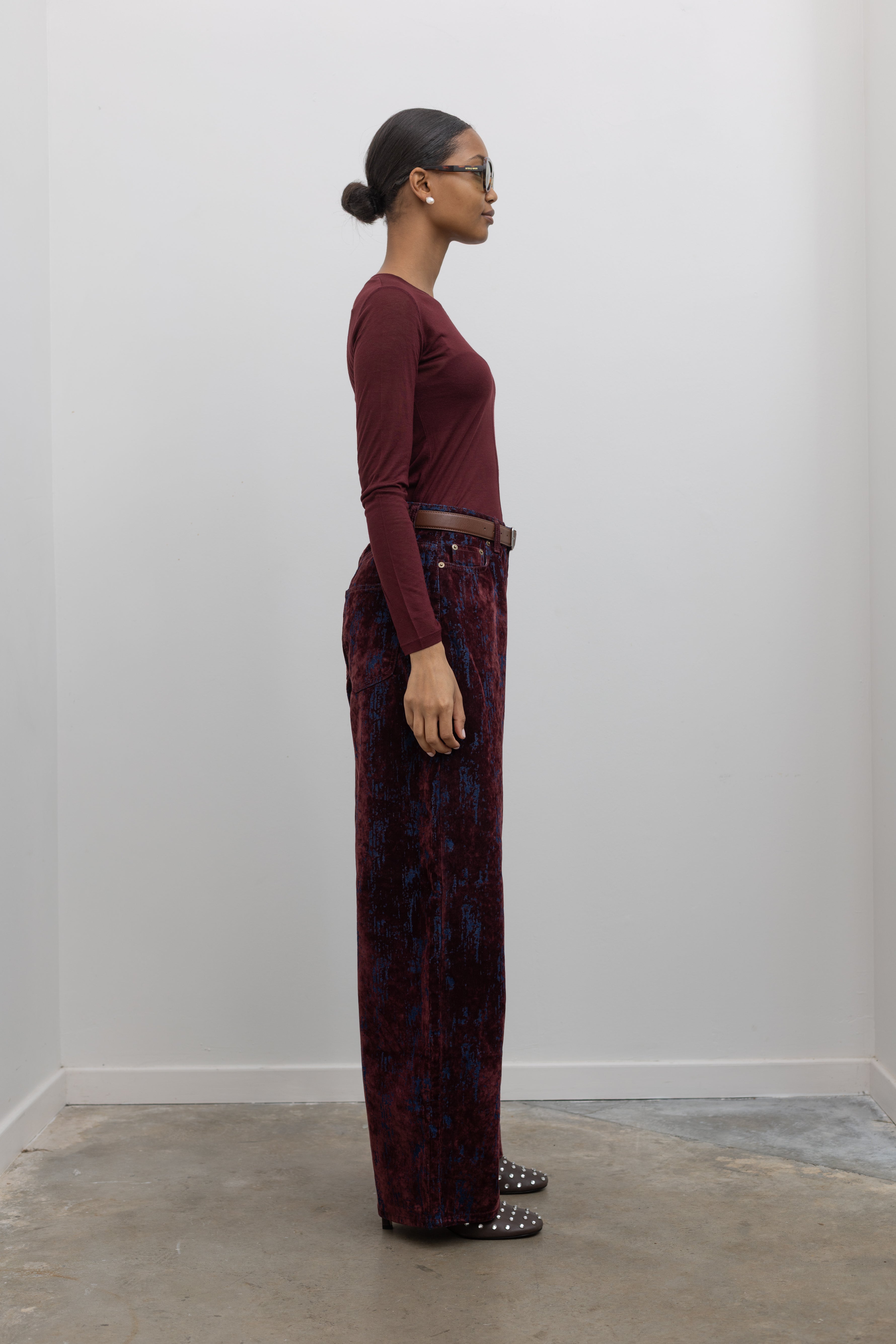 Full length side view of statement Paper Moon wide leg jeans in a luxurious burgundy distressed crackle velvet finish and a relaxed fit at an Alas Eius store. 