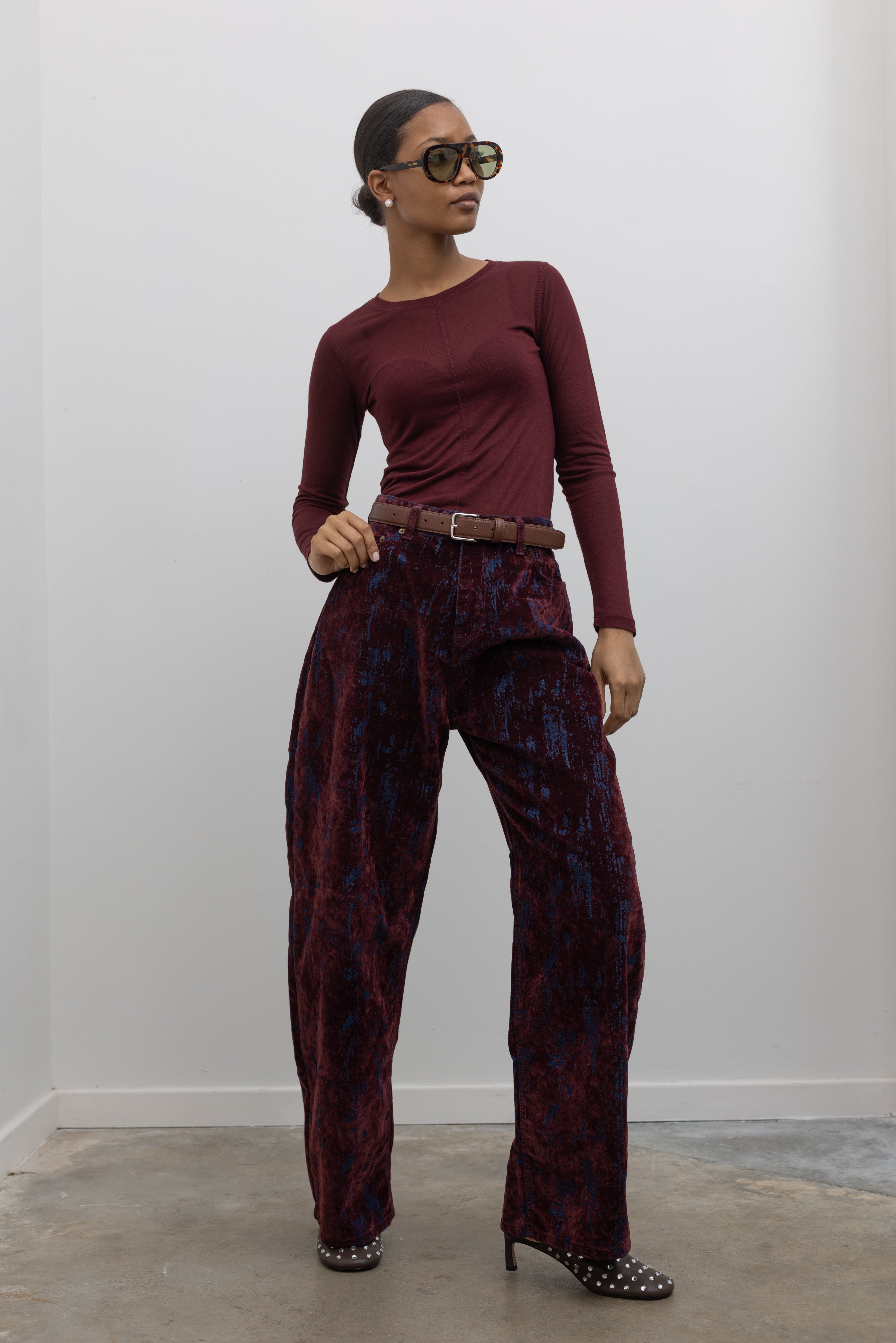 Full length front view of edgy yet chic Paper Moon wide leg jeans in a luxurious burgundy distressed crackle velvet finish and a relaxed fit paired with a burgundy fitted knit top and heels at an Alas Eius store. 