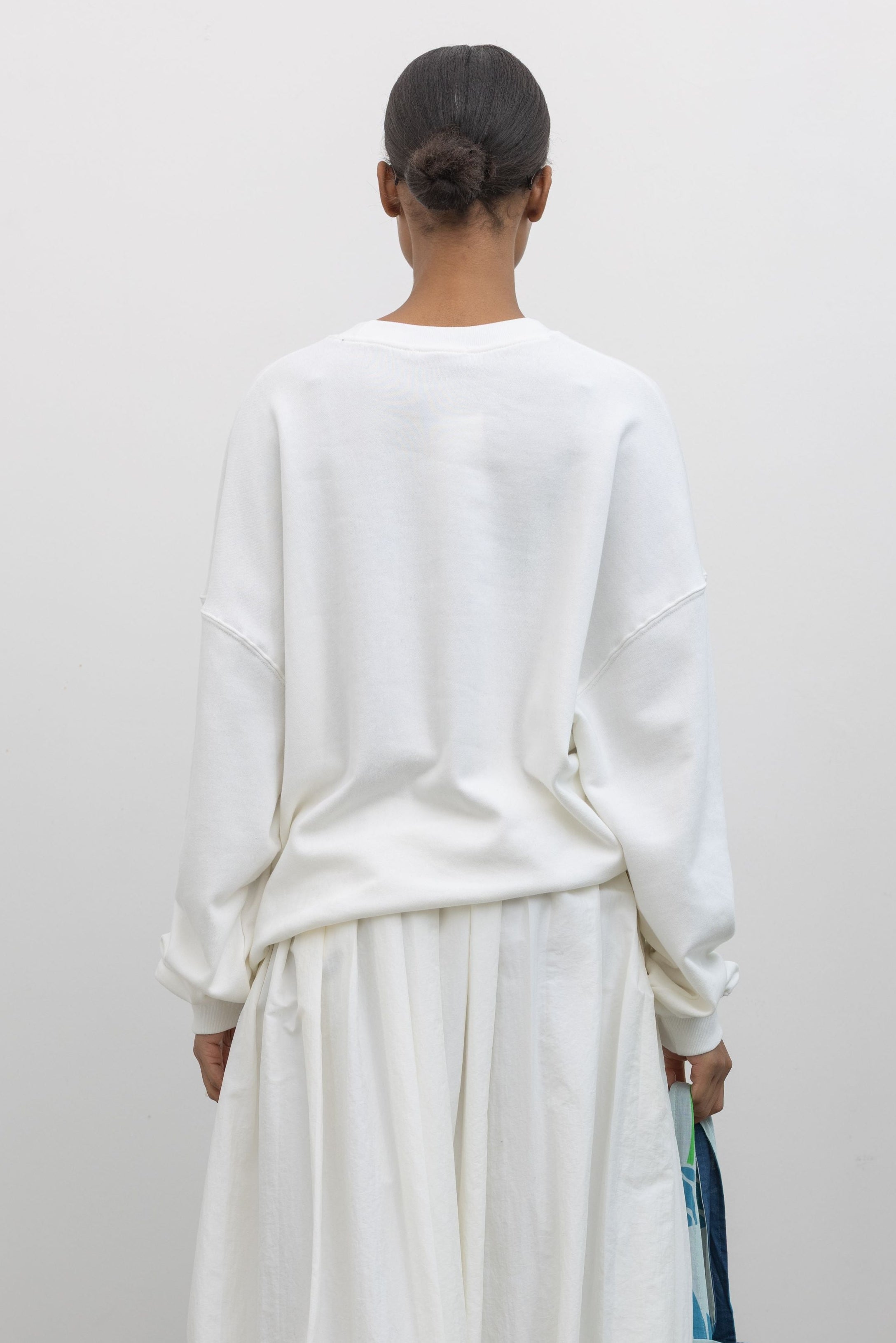 Back view of a woman wearing an oversized white crewneck sweatshirt with New York embroidered on the front in bright colors at an Alas Eius store. 
