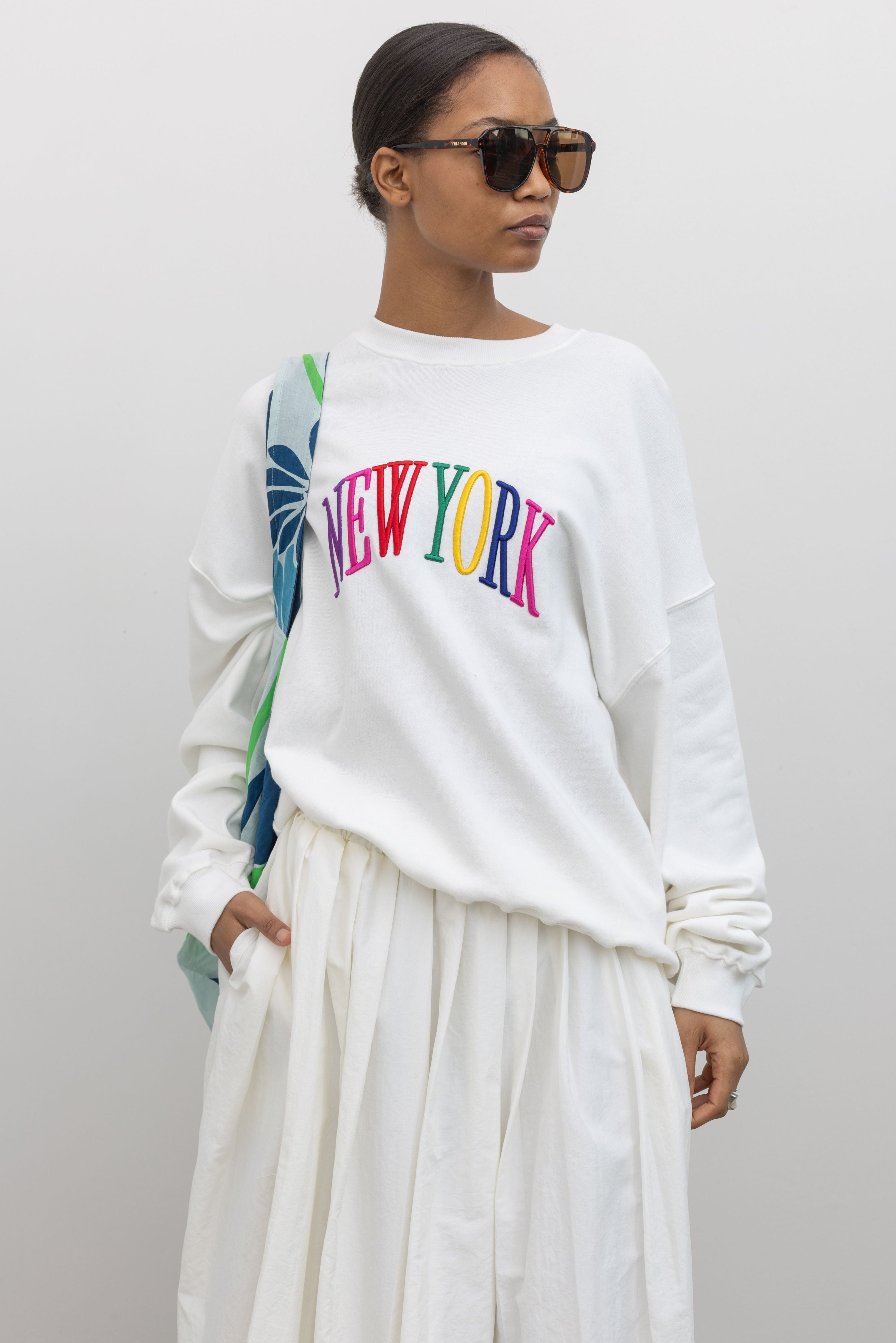 Woman standing wearing an oversized white crewneck sweatshirt with New York embroidered on the front in bright colors at Alas Eius. 