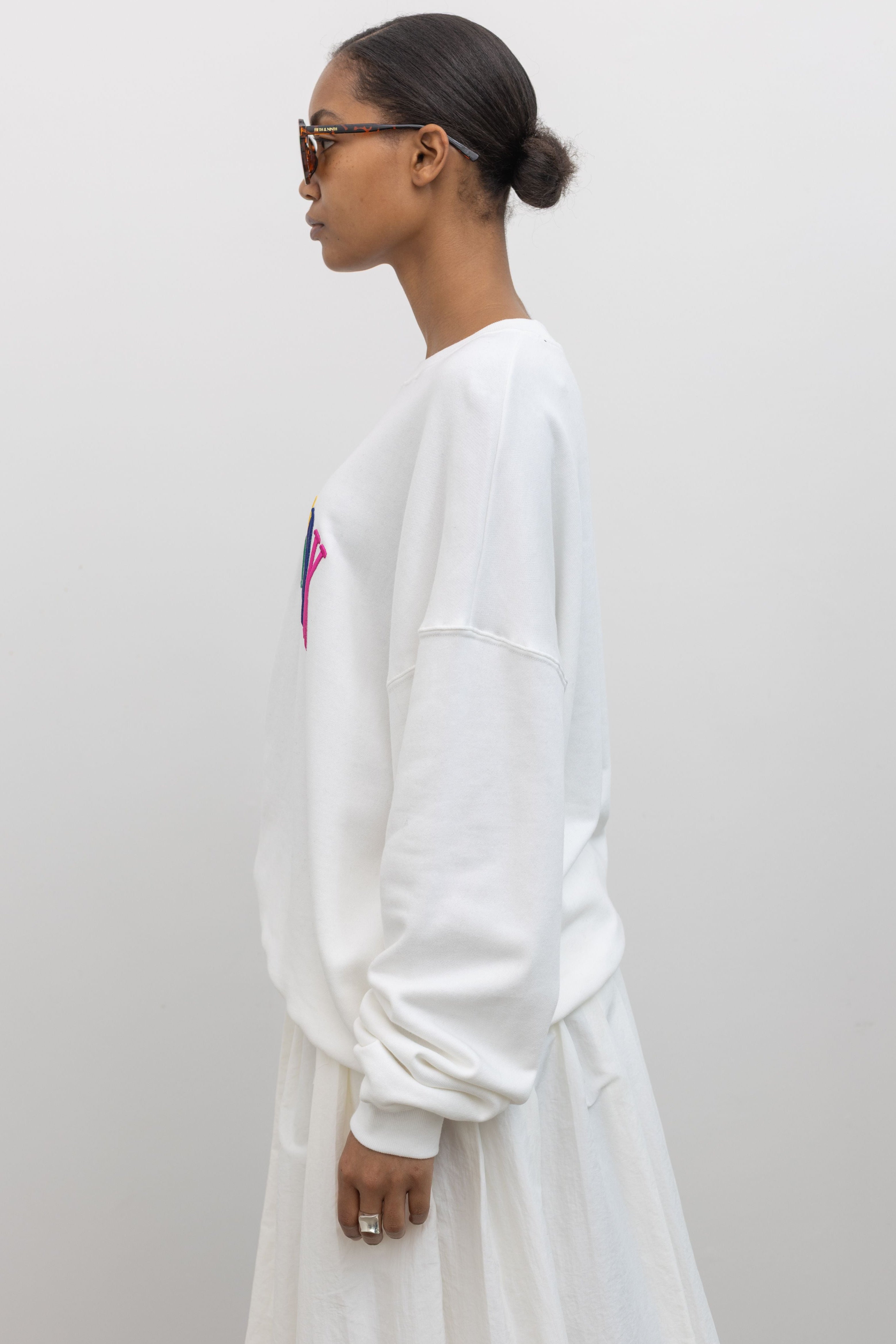 Side view of a woman wearing an oversized white crewneck sweatshirt with New York embroidered on the front in bright colors at an Alas Eius store. 
