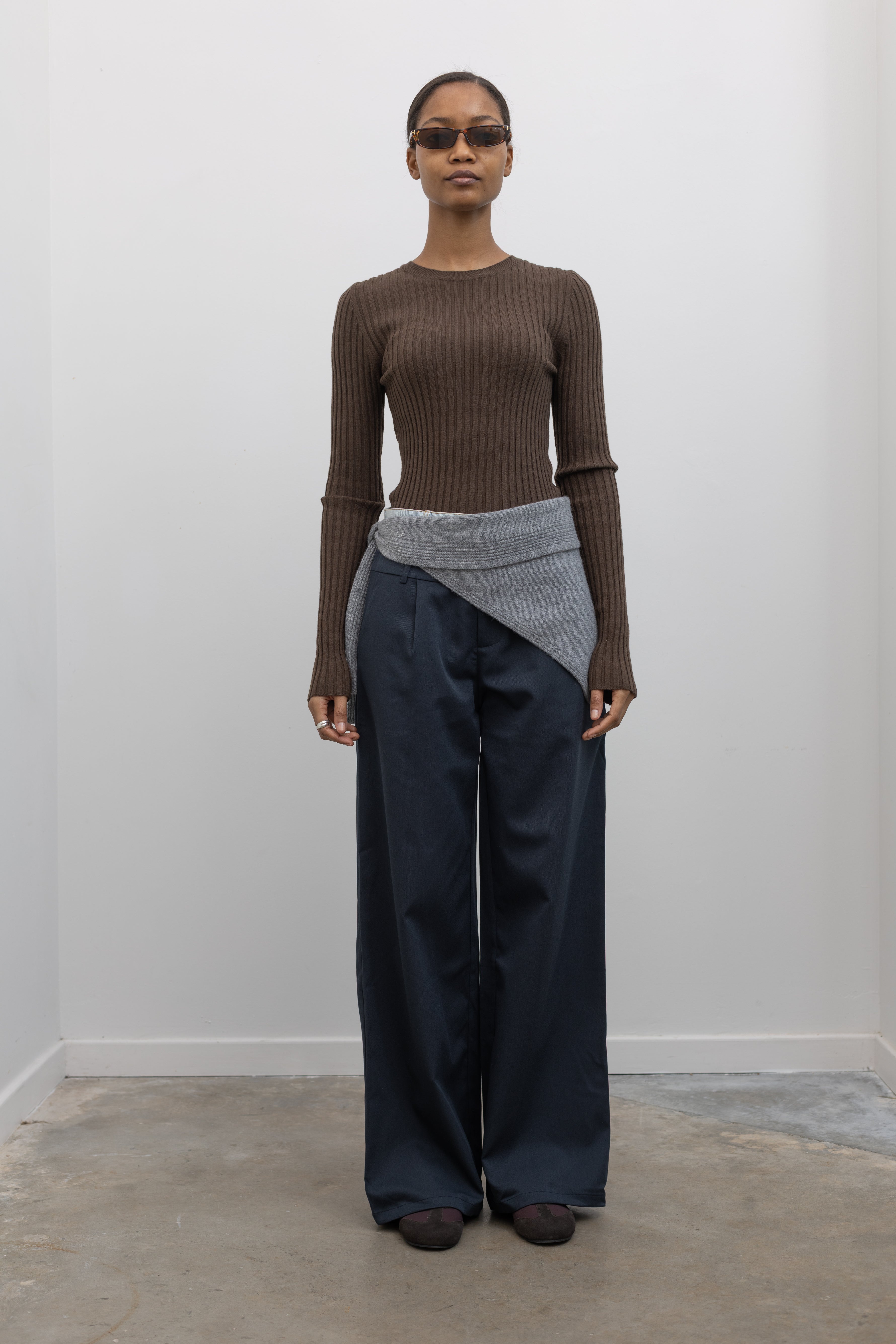 Full length view of a woman standing wearing spring fashion in a Paper Moon brand brown wool blend ribbed long sleeve fitted top with navy blue trousers and a gray bandana scarf around her waist at an Alas Eius store. 