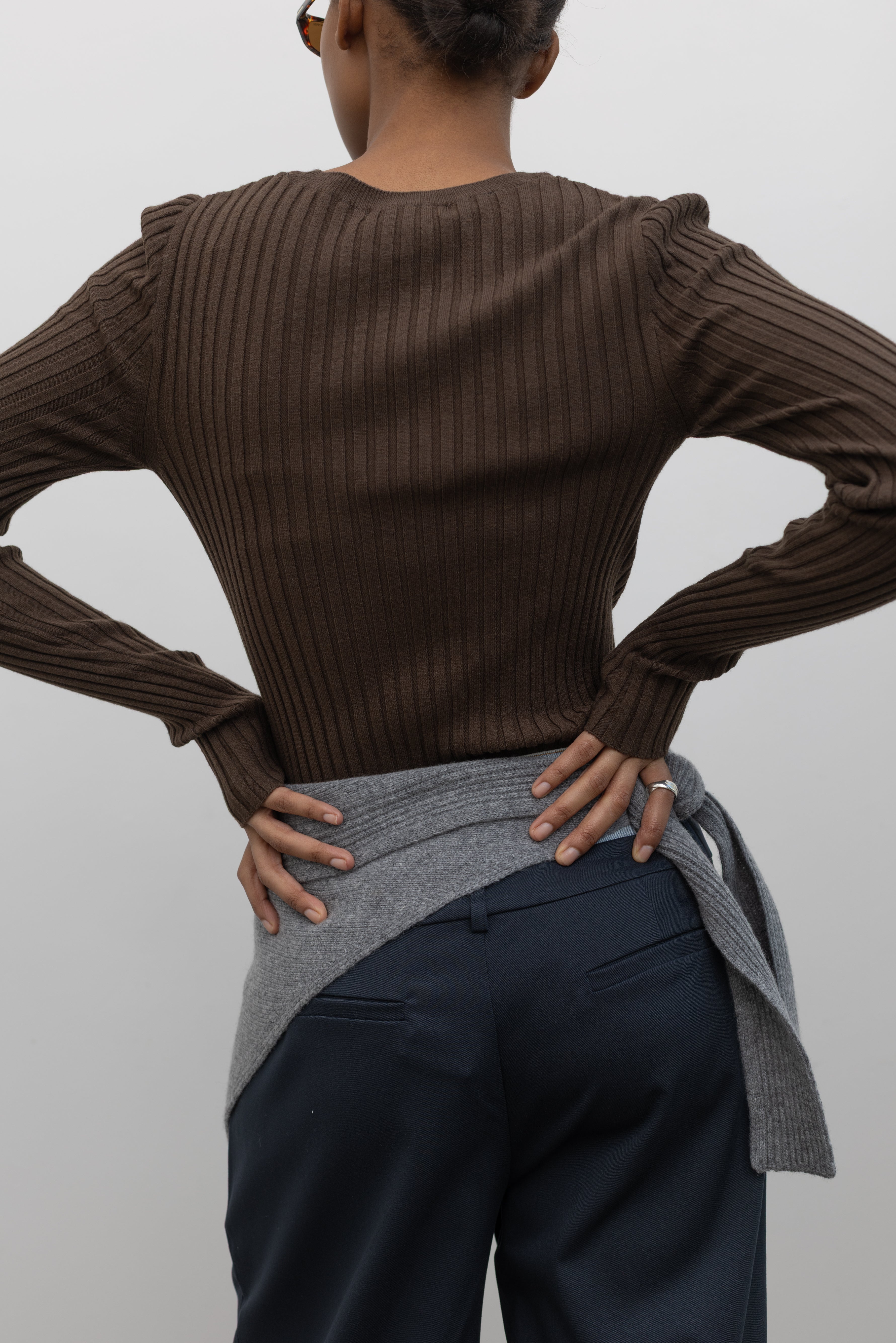 Close-up back view of a woman standing wearing an elevated essential spring staple in a Paper Moon brand brown wool blend ribbed long sleeve fitted top with navy blue trousers and a gray bandana scarf around her waist at an Alas Eius 