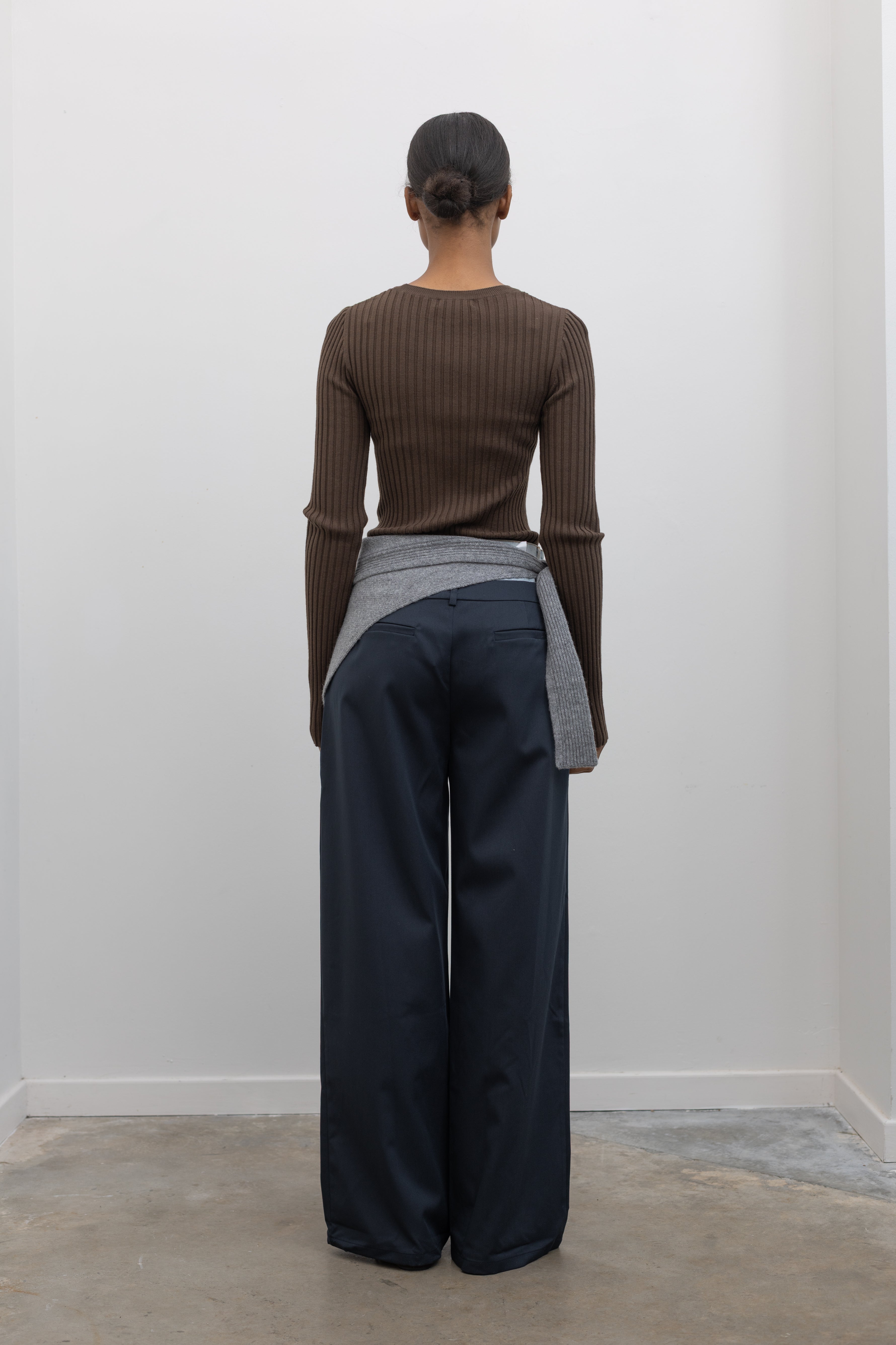 Full length back view of a woman standing wearing an elevated essential spring staple in a Paper Moon brand brown wool blend ribbed long sleeve fitted top with navy blue trousers and a gray bandana scarf around her waist at an Alas Eius 