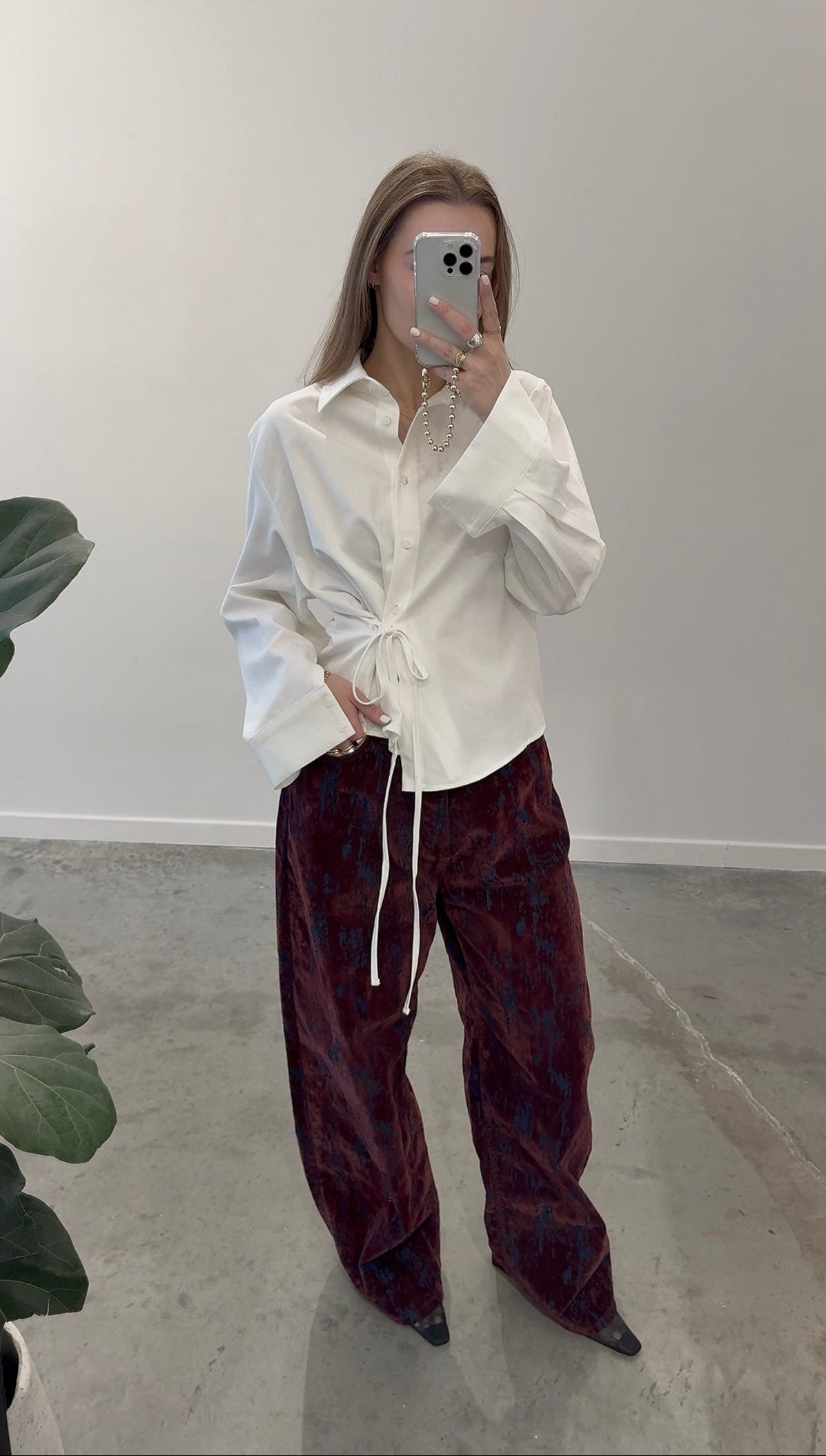 Full length front view of a woman wearing unique Paper Moon wide leg jeans in a luxurious burgundy distressed crackle velvet finish and an Alas Eius label side tie button down shirt at an Alas Eius store. 