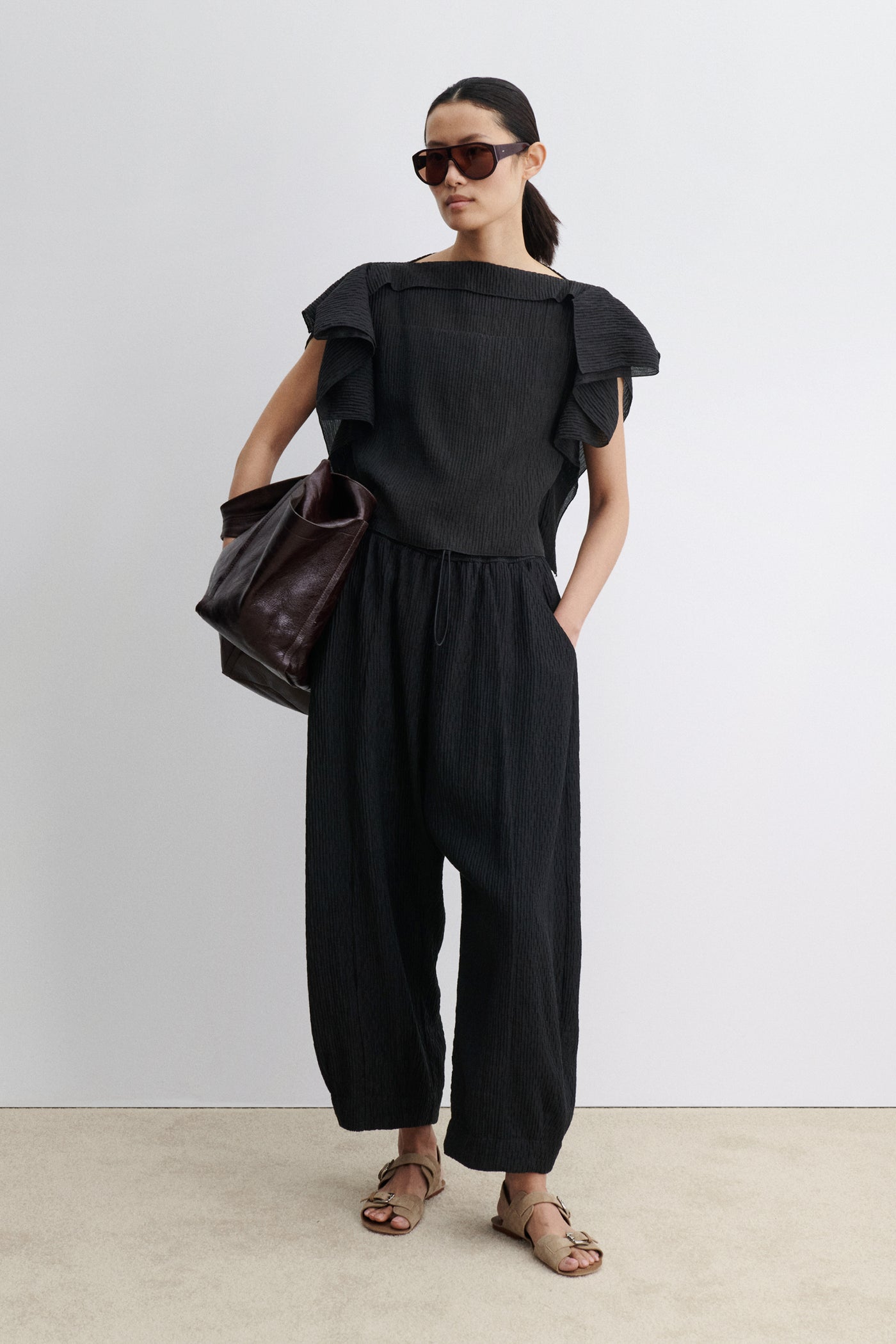 Full length front view of a woman standing wearing contemporary spring and summer womenswear fashion in a coordinating set by Rachel Comey. The  airy Black Rivera Top features softly crinkled fabric, cascading flutter sleeves, and a relaxed, boxy silhouette. Perfectly paired with coordinating Ashbolt Pant for a cohesive look at an Alas Eius Store. Made in the USA. 