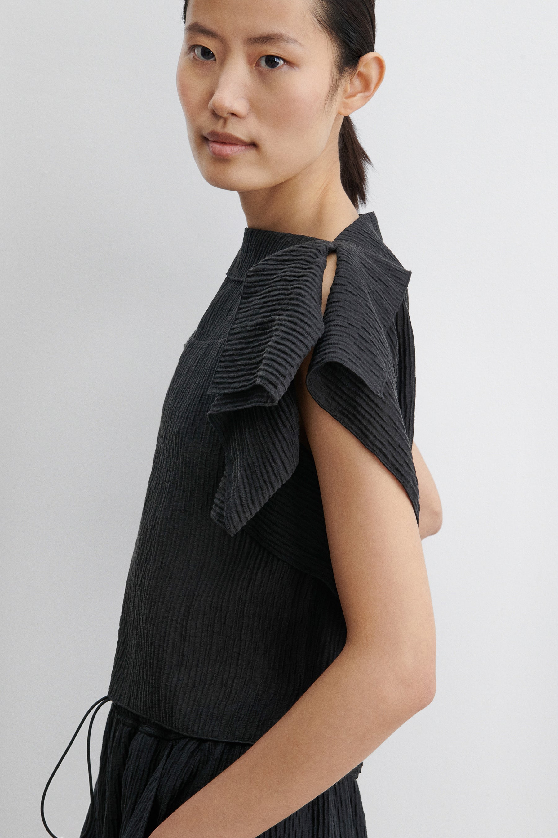 Close-up side view of a woman standing wearing contemporary spring and summer womenswear fashion in a coordinating set by Rachel Comey. The airy Black Rivera Top features softly crinkled fabric, cascading flutter sleeves, and a relaxed, boxy silhouette. Perfectly paired with coordinating Ashbolt Pant for a cohesive look at an Alas Eius Store. Made in the USA. 