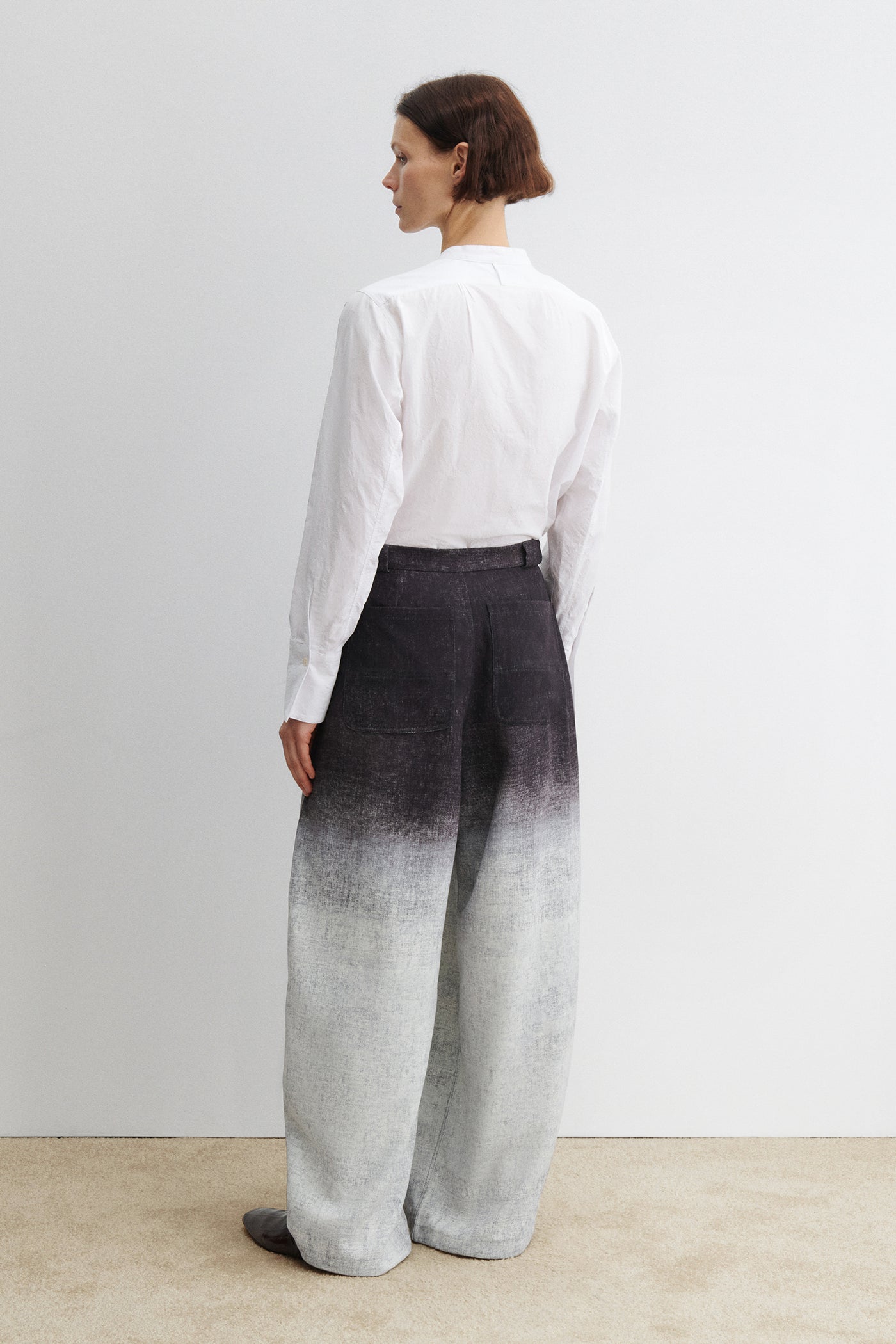 Full length back view of a woman standing wearing spring fashion in the Rachel Comey black Dias pant featuring a high waist and wide-leg silhouette, cut from softly textured fabric with a natural drape and ombre effect. Front pleats add volume and ease, creating a relaxed fit through the leg at Alas Eius.