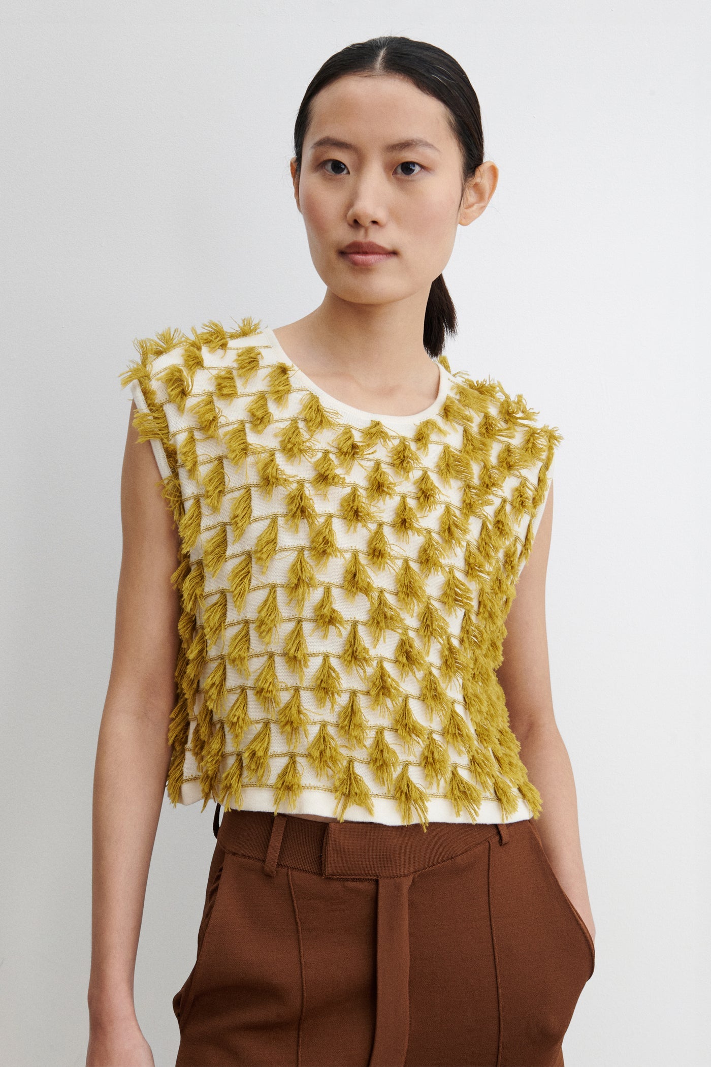 Close-up view of a woman posing wearing retro and vintage inspired fashion in a Rachel Comey Cotton and Baby Alpaca Cream Mustard Hazlitt sleeveless knit top detailed with rows of playful, three-dimensional fringes. Designed with a relaxed, boxy cut and finished with a soft crew neckline at Alas Eius. Made in Peru. 