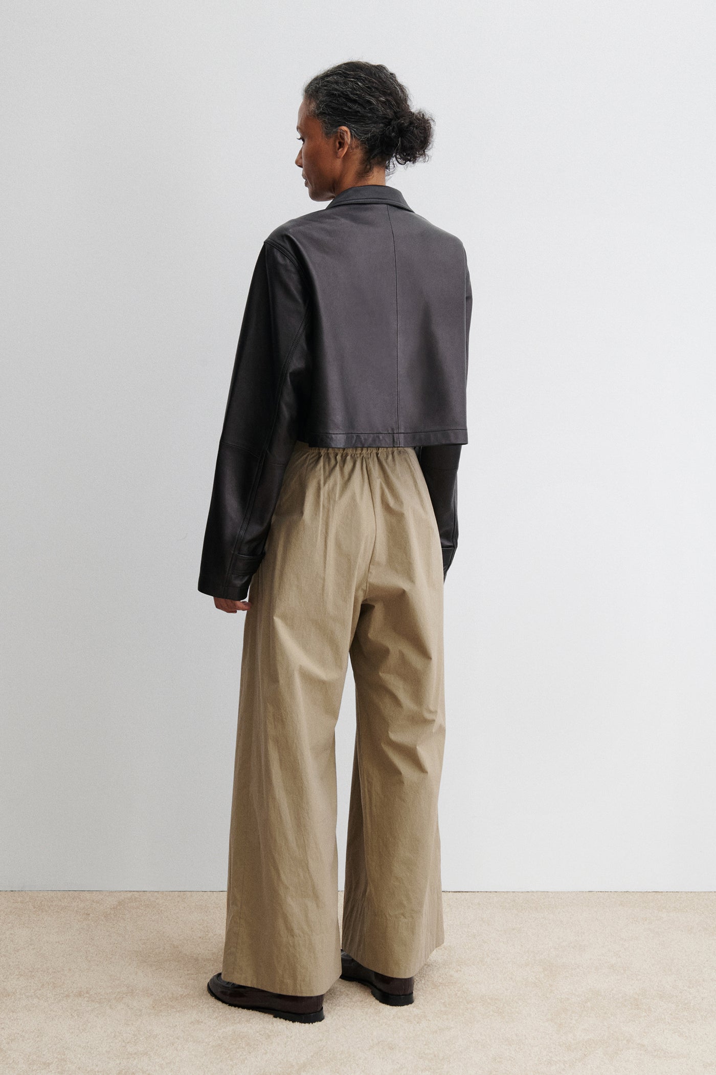 Full length back view of a woman standing wearing chic and versatile spring summer fashion in the Rachel Comey Khaki Coxsone Pant with front pleats, high-waist, and wide leg, cut from signature Organic Crisp cotton. The trouser features side slip pockets, an elastic waistband at the back, a concealed side zipper paired with a cropped faux leather button front jacket at an Alas Eius store. Made in the USA.