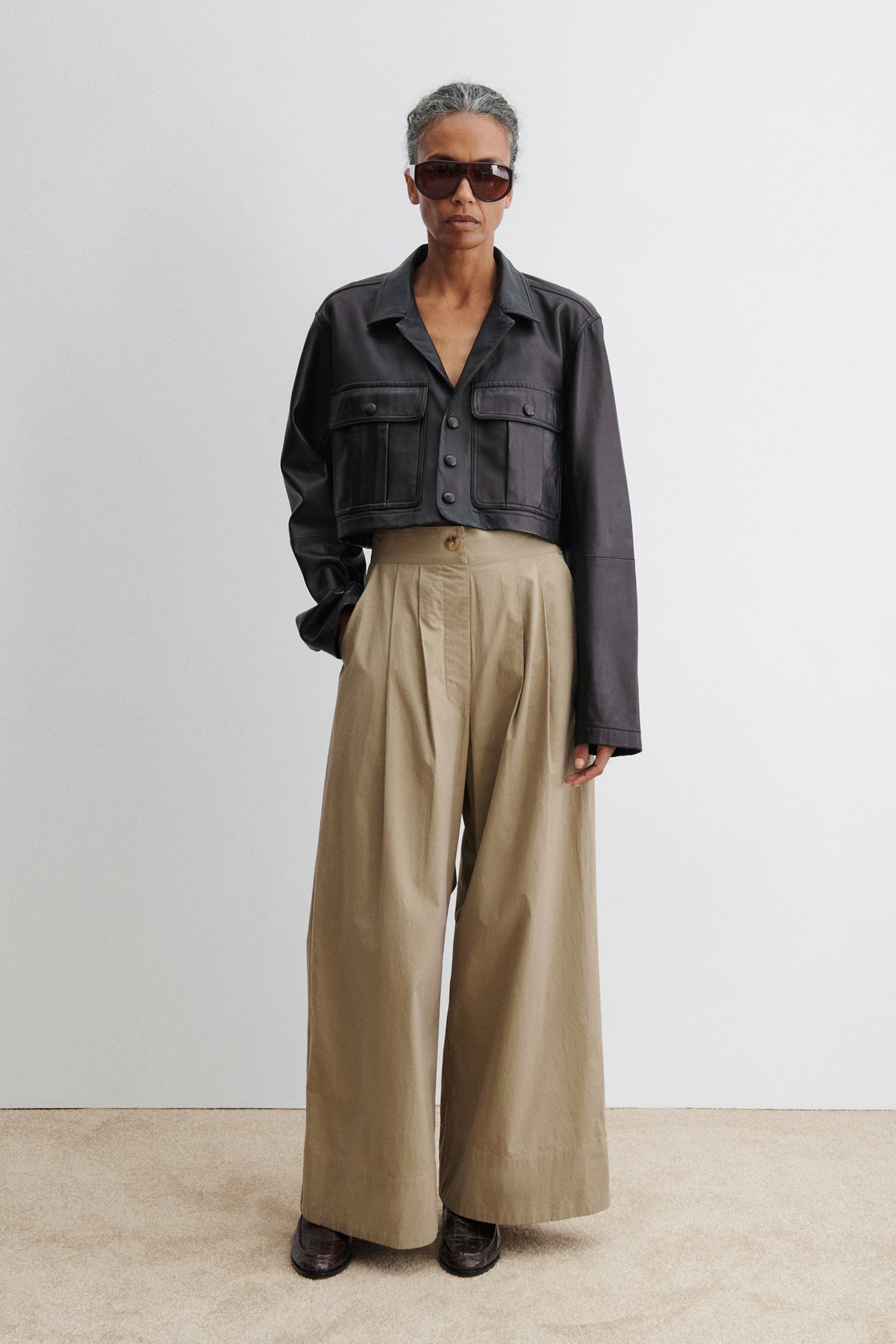 Woman standing wearing chic and versatile spring summer fashion in the Rachel Comey Khaki Coxsone Pant with front pleats, high-waist, and wide leg, cut from signature Organic Crisp cotton. The trouser features side slip pockets, an elastic waistband at the back, a concealed side zipper paired with a cropped faux leather button front jacket at an Alas Eius store. Made in the USA. 