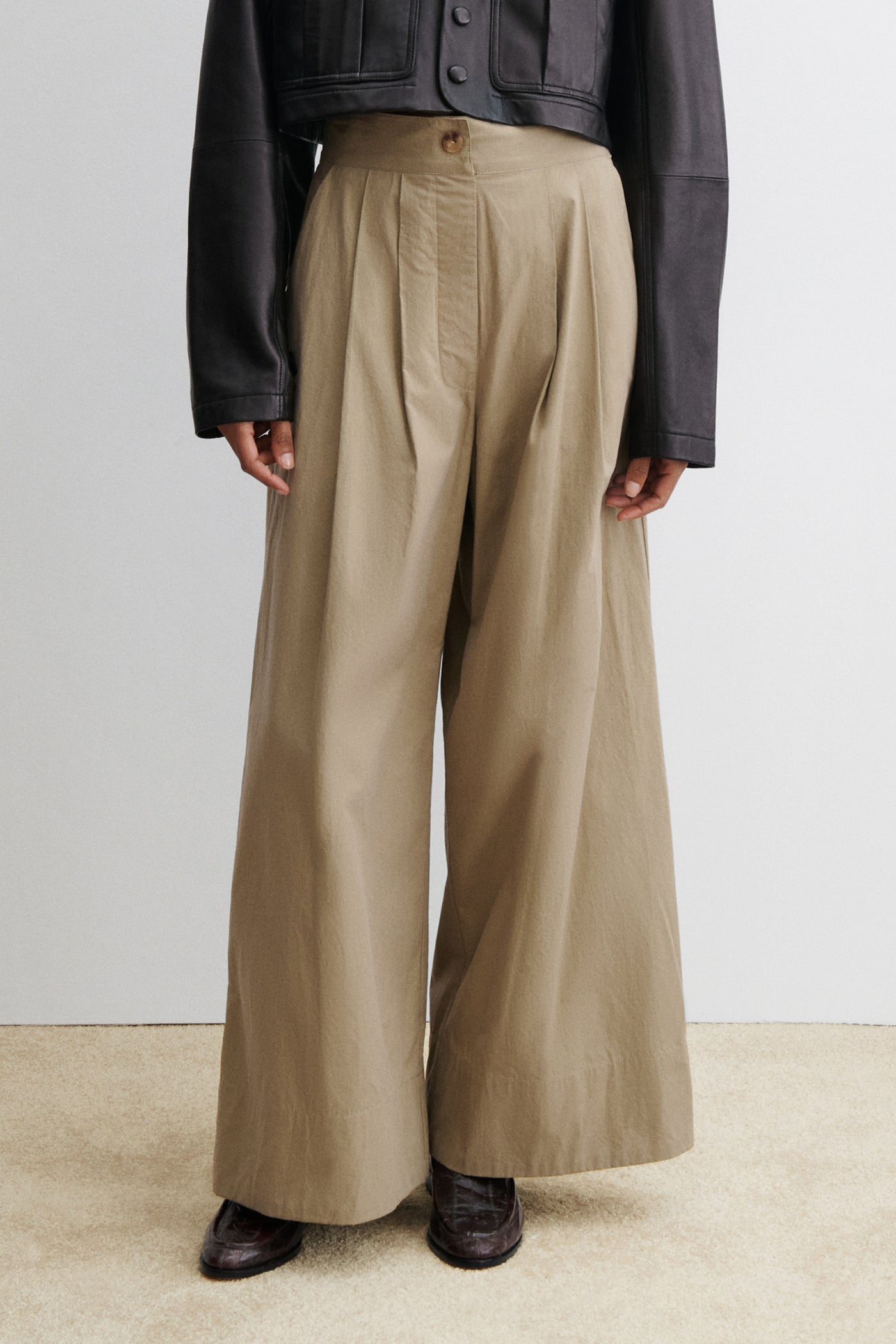 Woman standing wearing chic and versatile spring summer fashion in the Rachel Comey Khaki Coxsone Pant with front pleats, high-waist, and wide leg, cut from signature Organic Crisp cotton. The trouser features side slip pockets, an elastic waistband at the back, a concealed side zipper paired with a cropped faux leather button front jacket at an Alas Eius store. Made in the USA. 