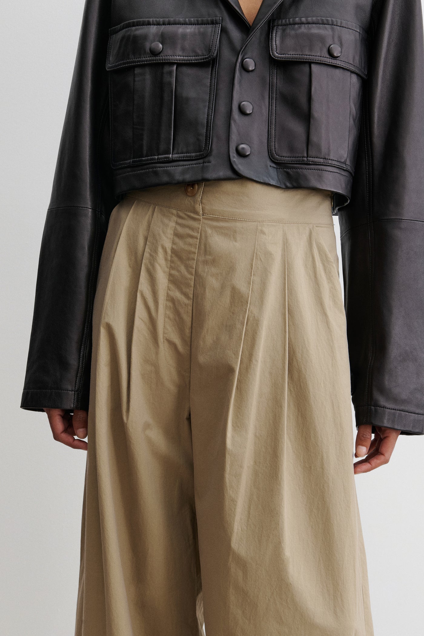 Close-up detail view of a woman standing wearing chic and versatile spring summer fashion in the Rachel Comey Khaki Coxsone Pant with front pleats, high-waist, and wide leg, cut from signature Organic Crisp cotton. The trouser features side slip pockets, an elastic waistband at the back, a concealed side zipper paired with a cropped faux leather button front jacket at an Alas Eius store. Made in the USA. 